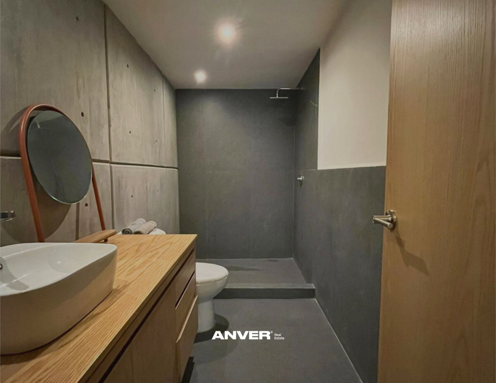 Baño moderno con lavabo de madera, espejo circular, inodoro, paredes de concreto y baldosas grises, ducha sin puertas y puerta de madera.