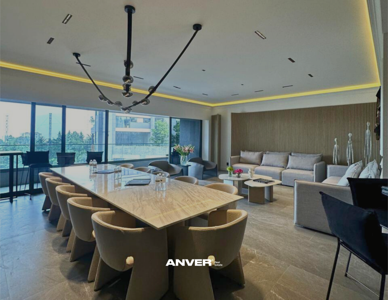 Sala de reuniones moderna con mesa de madera, sillas beige, sofá gris, mesas auxiliares, decoración con estatuas y plantas, amplio ventanal con vista a edificios y árboles, iluminación moderna, logotipo de ANVER en la parte inferior