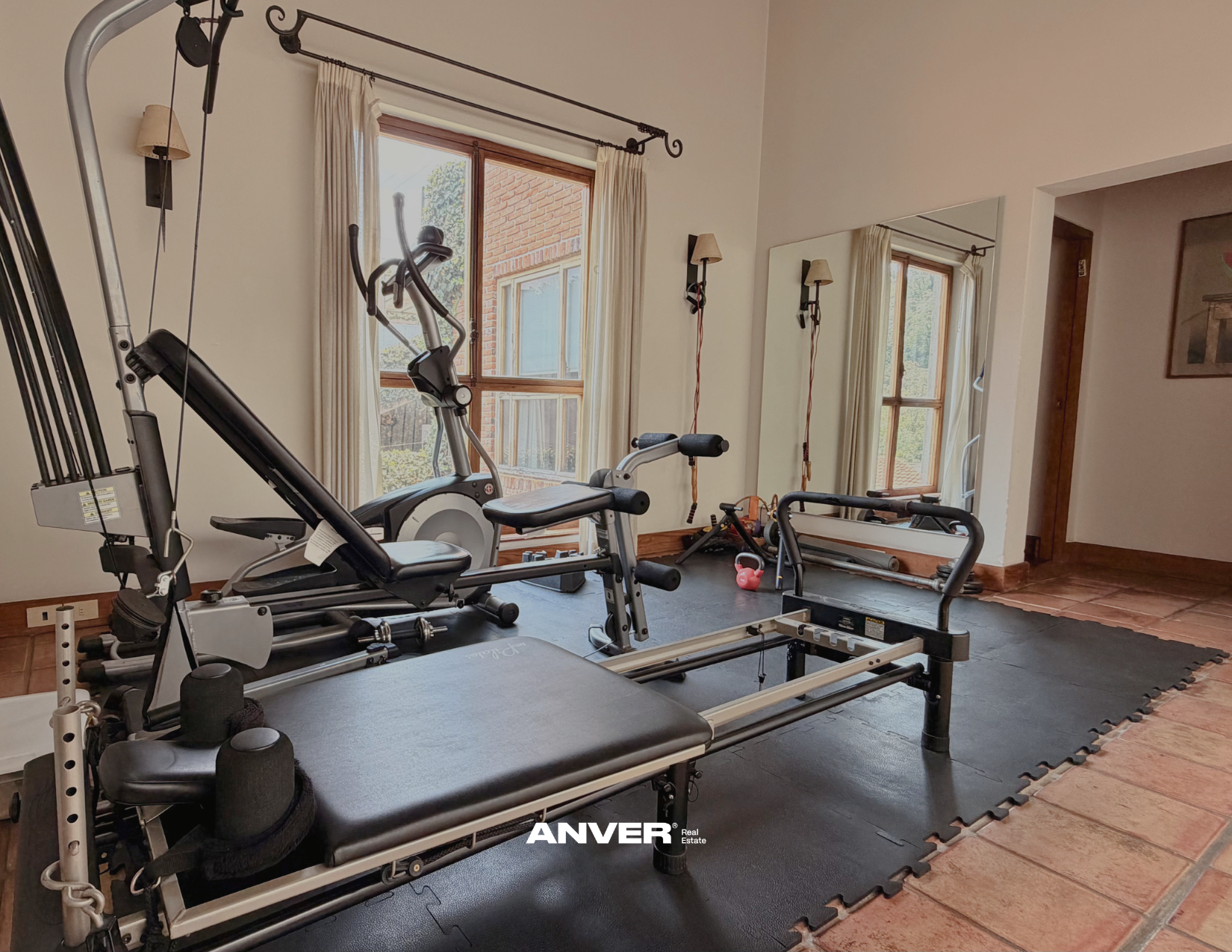 Sala de entrenamiento en casa con equipo de ejercicio, including una máquina de remo, bicicleta fija, esteras, pesas y kettlebell, con ventanas que dejan entrar luz natural y paredes decoradas con espejos y lámparas de pared.