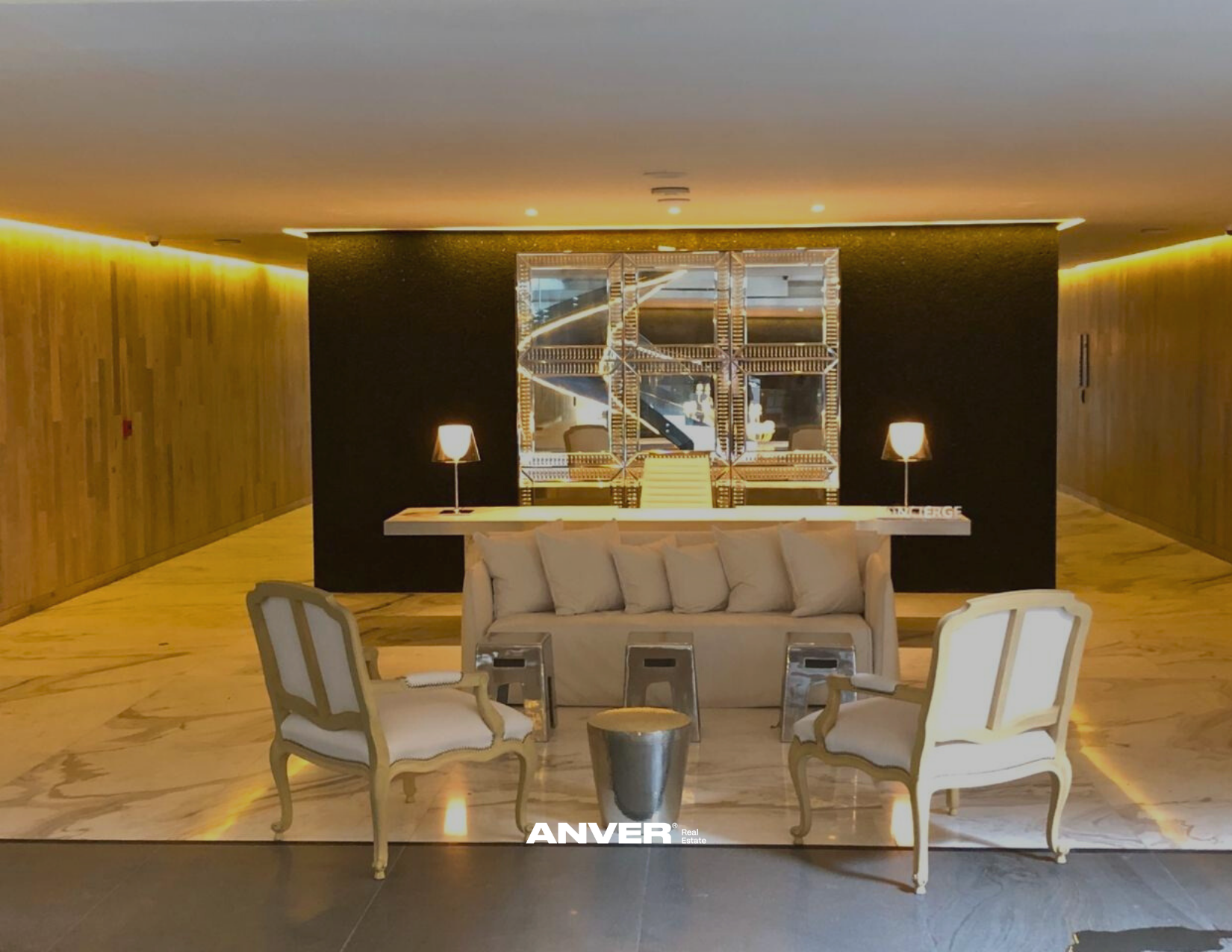 Sala de lobby con sillones blancos y mesas de centro, fondo con espejo y lámparas, paredes de madera y suelo de mármol.