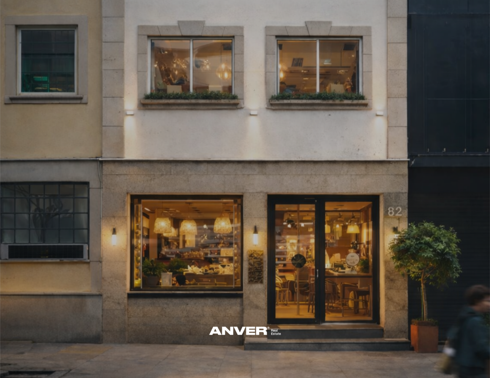 Fachada de un restaurante o cafetería con gran ventanal iluminado, plantas y detalles arquitectónicos en la fachada.