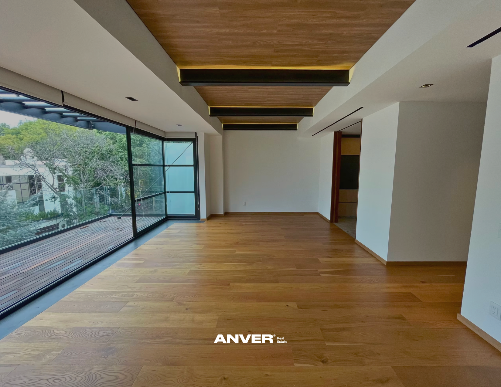 Espacio interior moderno con suelos de madera, paredes blancas y ventana de piso a techo con vista a un balcón y árboles afuera. Techo con elementos de madera y luces empotradas.