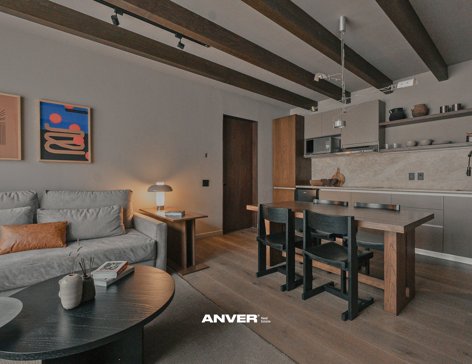 Interior de una sala de estar y cocina moderna con paredes grises, muebles de madera y decoraciones minimalistas. Incluye sofá gris con cojín marrón, mesa de noche con lámpara y libros, y comedor con mesa de madera y sillas negras. La cocina tiene gabinetes grises, estantes abiertos con utensilios y decoraciones, y contraseñas de piedra.