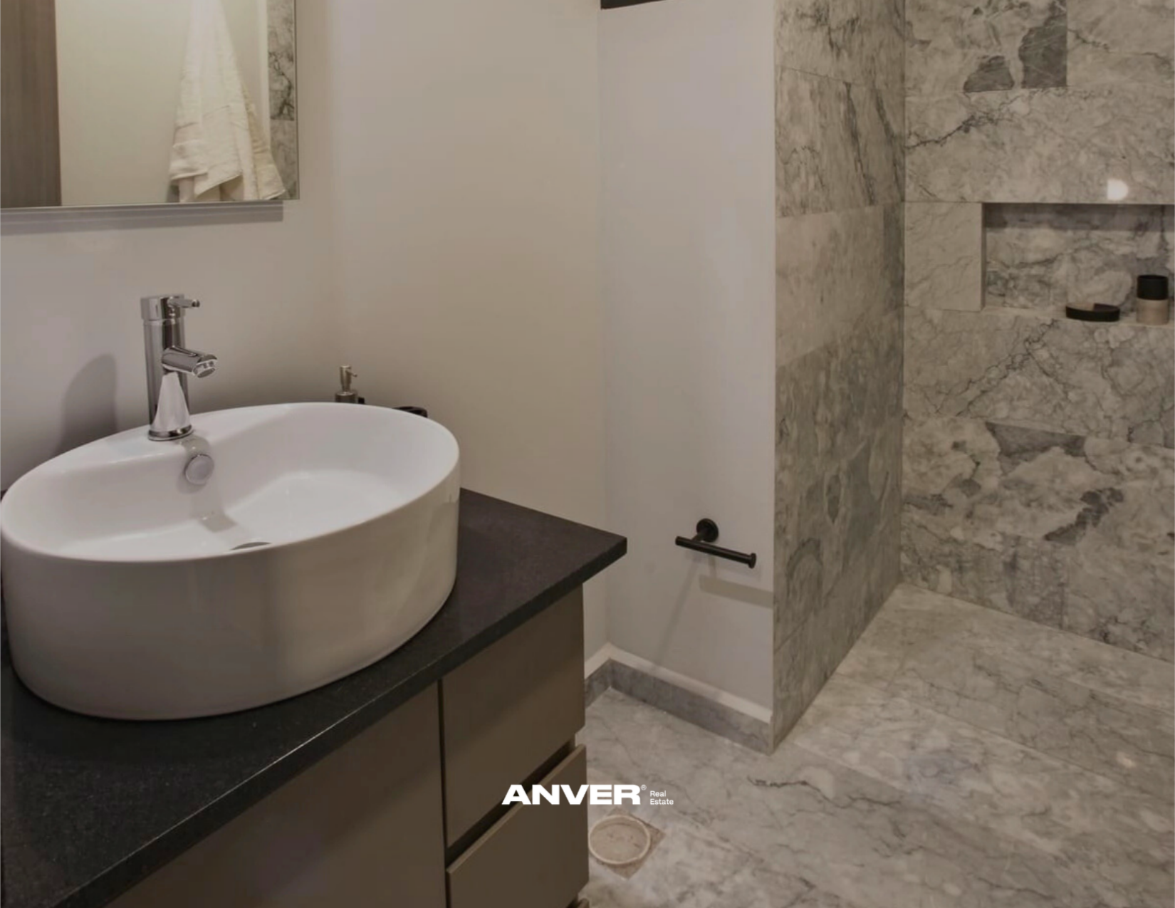 Lavabo de cerámica sobre mueble oscuro, espejo, toalla en la pared, área de ducha con paredes de piedra y un estante con objetos, suelo de mármol.
