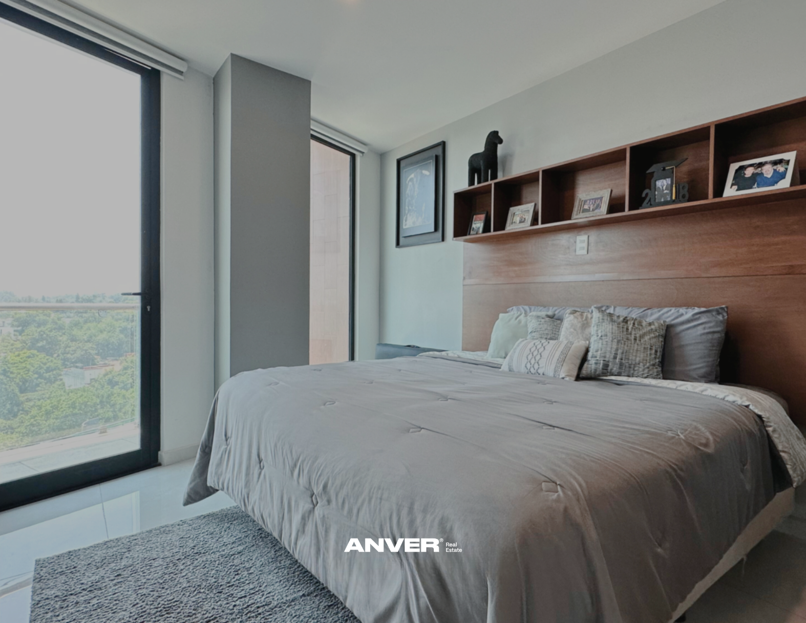 Habitación moderna con cama de matrimonio, almohadas decorativas, ventanales con vista a árboles y ciudad, estantería con marcos de fotos y decoraciones, paredes blancas y piso de cerámica.
