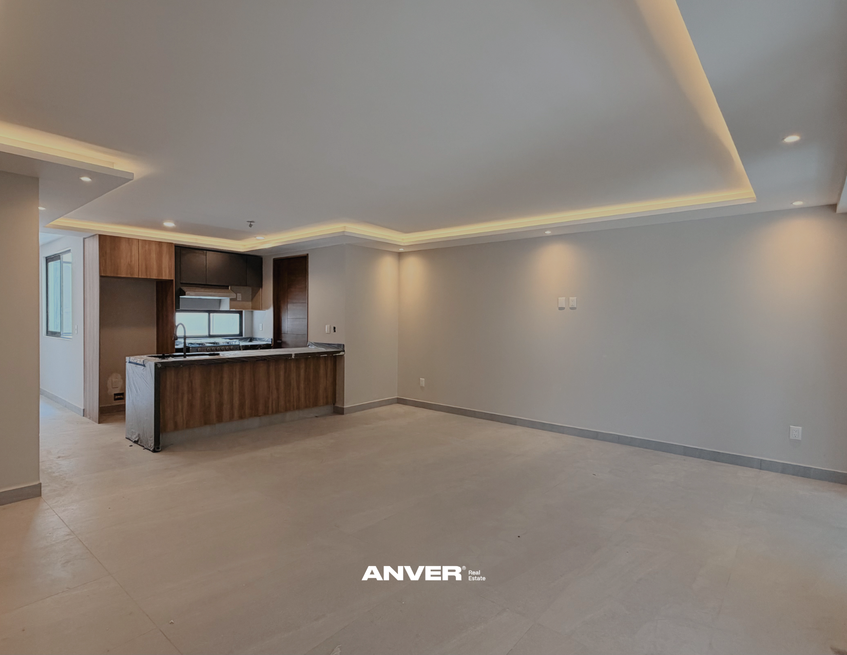 Espacioso salón abierto con cocina de madera y encimeras de mármol, paredes blancas, acabado de techo con iluminación empotrada en yeso, ventanas grandes y piso de baldosas claras.