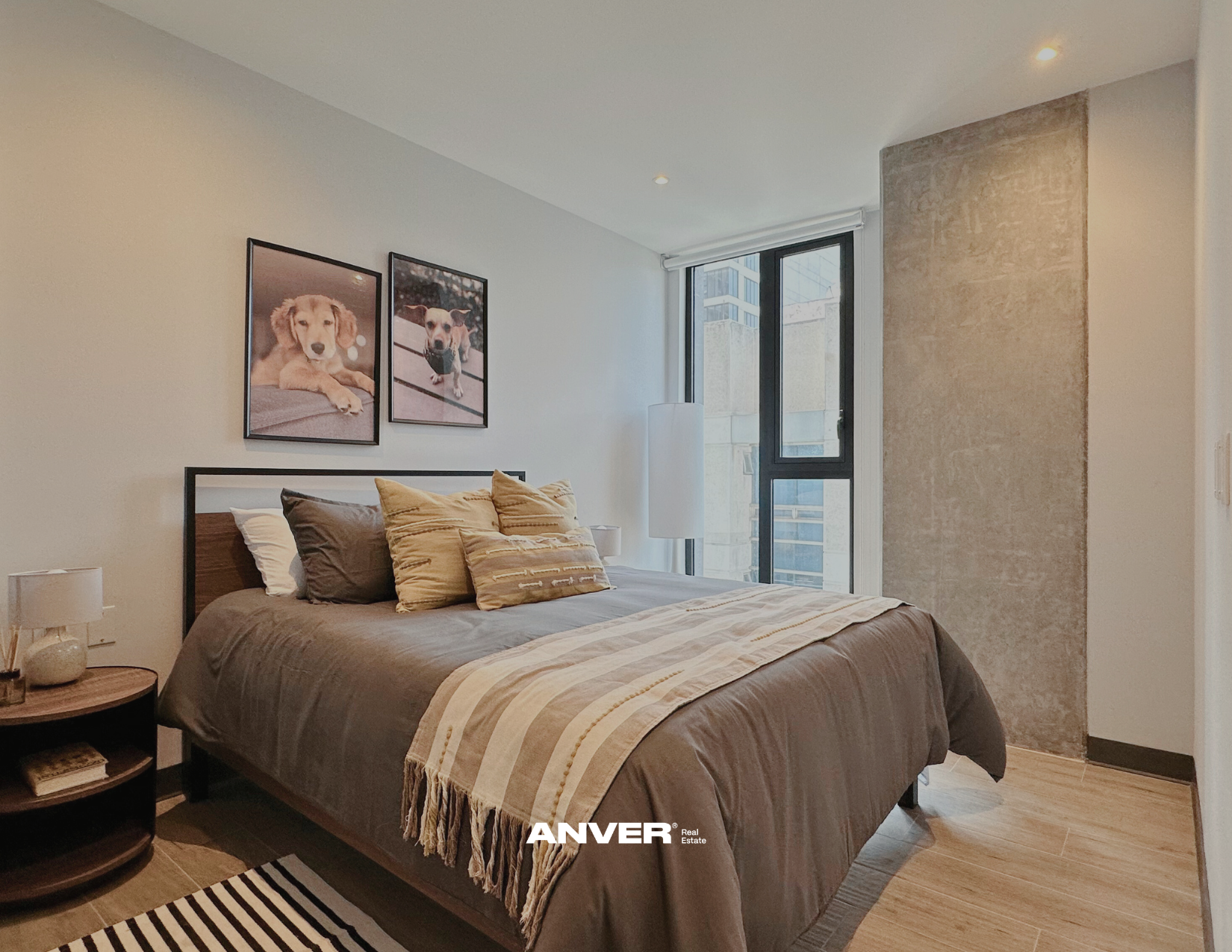 Dormitorio con cama de sábanas grises y cojines beige, una lámpara de piso junto a la ventana y cuadros de perros en la pared. La habitación tiene piso de madera clara y una pared con acabado de concreto. La ventana grande permite la entrada de luz natural.