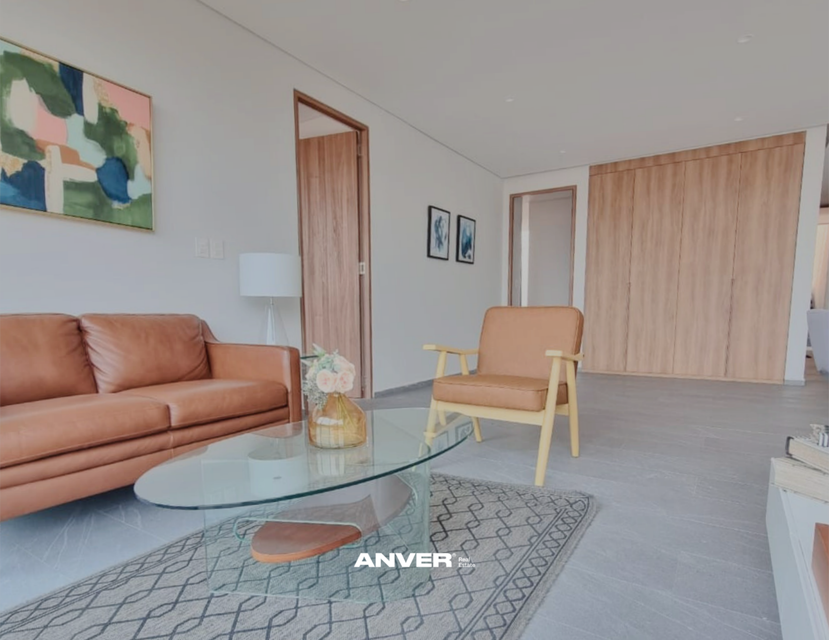 Sala moderna con sofá de cuero marrón, silla amarilla de madera, mesa de vidrio con florero, pared con cuadros decorativos, puertas de madera y alfombra. 
