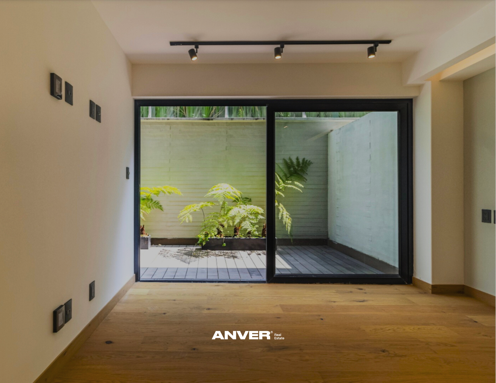 Vista del interior de una sala con puertas de cristal que conducen a un pequeño patio con plantas y un muro blanco.
