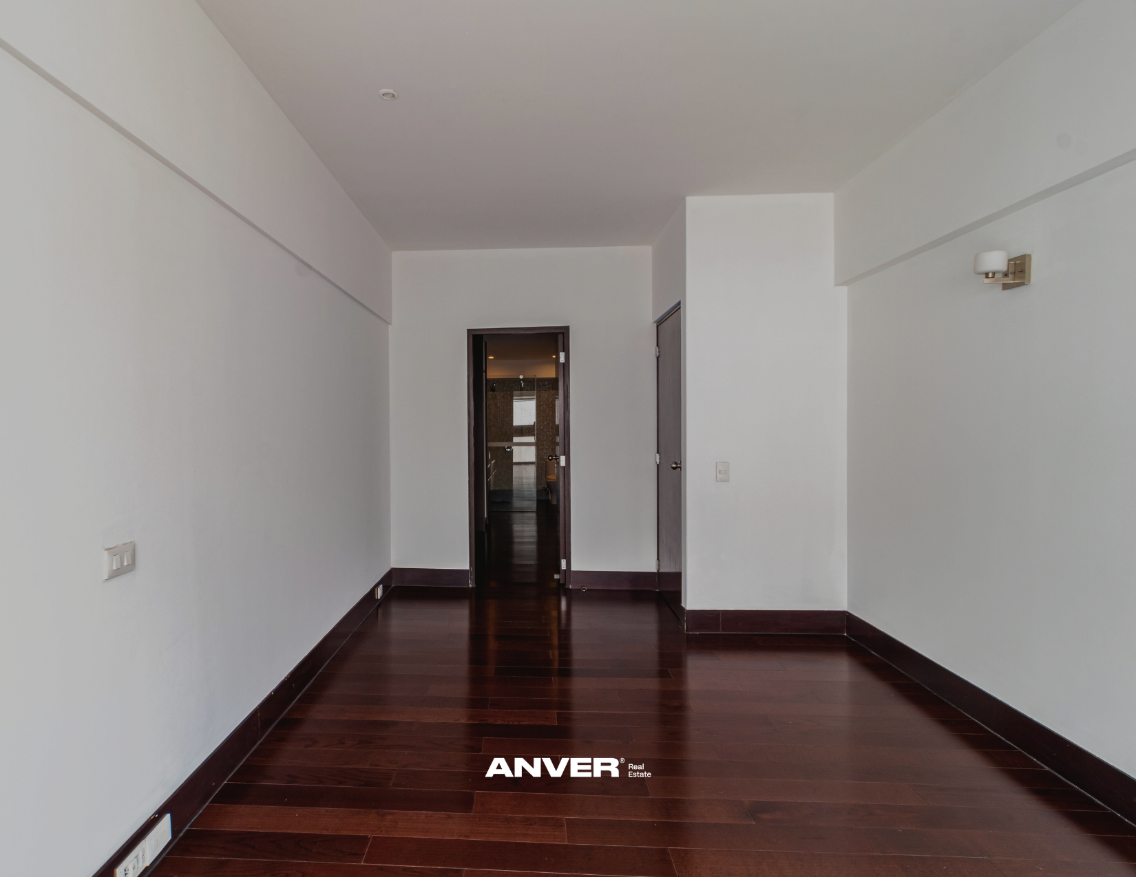 Habitación vacía con paredes blancas, suelo de madera oscura, y puertas en el fondo, con un sutil decorado minimalista.