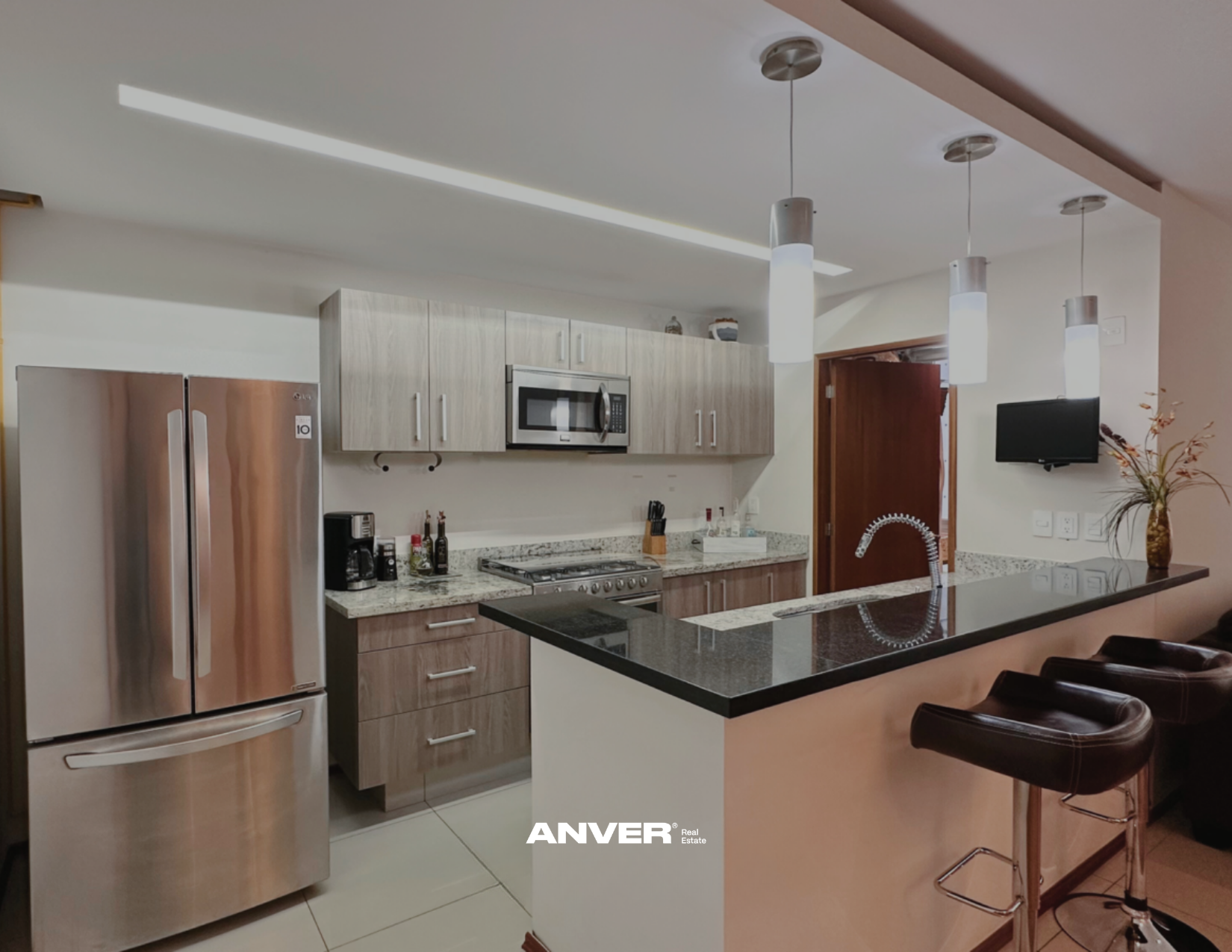 Cocina moderna con refrigerador de doble puerta, microondas empotrado, estufa, encimera de granito y barra con asientos. Decoración sencilla con un jarrón con plantas y televisión en la pared.