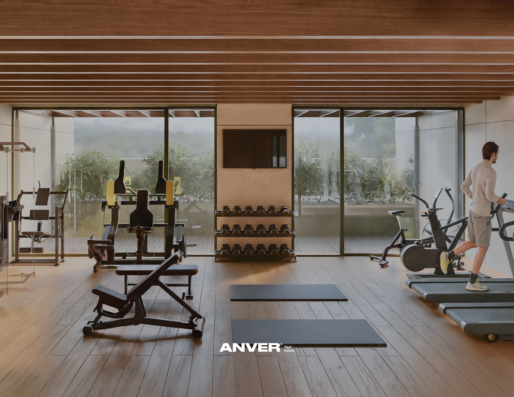 Interior de un gimnasio con pesas, máquinas de ejercicio y un hombre corriendo en una caminadora, con ventanas grandes que permiten la vista de árboles y naturaleza exterior.