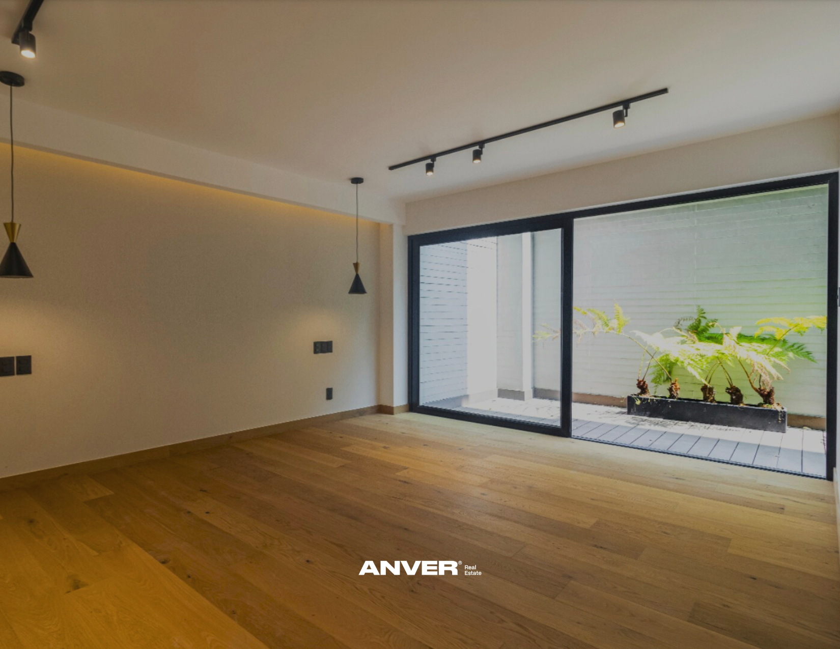 Interior de una habitación con paredes blancas, pisos de madera y un ventanal grande con vista a un patio con plantas verdes en una maceta larga.