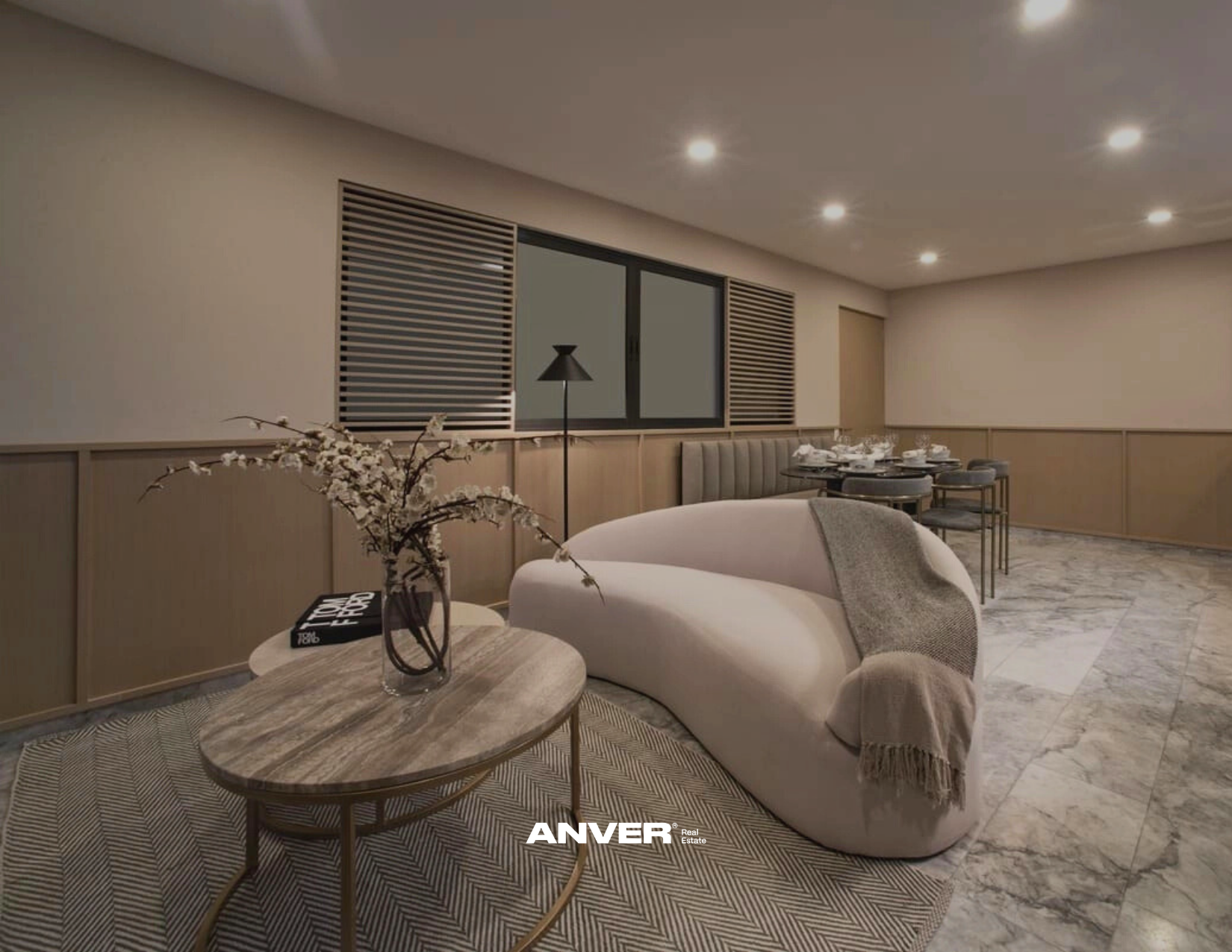 Sala de estar con sillón blanco, mesa de centro, flores, lámpara y comedor de vidrio y madera, en un ambiente elegante y moderno con paredes de madera y cerámica en el suelo.