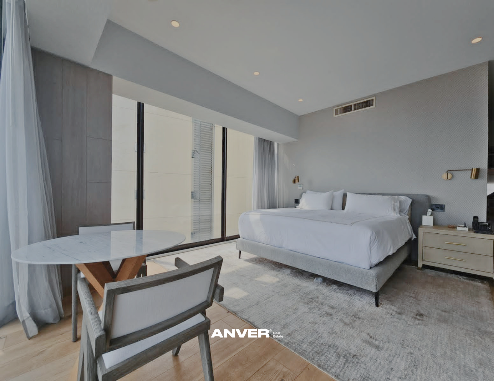 Habitación moderna con cama grande, paredes grises y cortinas blancas, vista al exterior a través de ventanas de piso a techo, mesa redonda y sillas en primer plano.