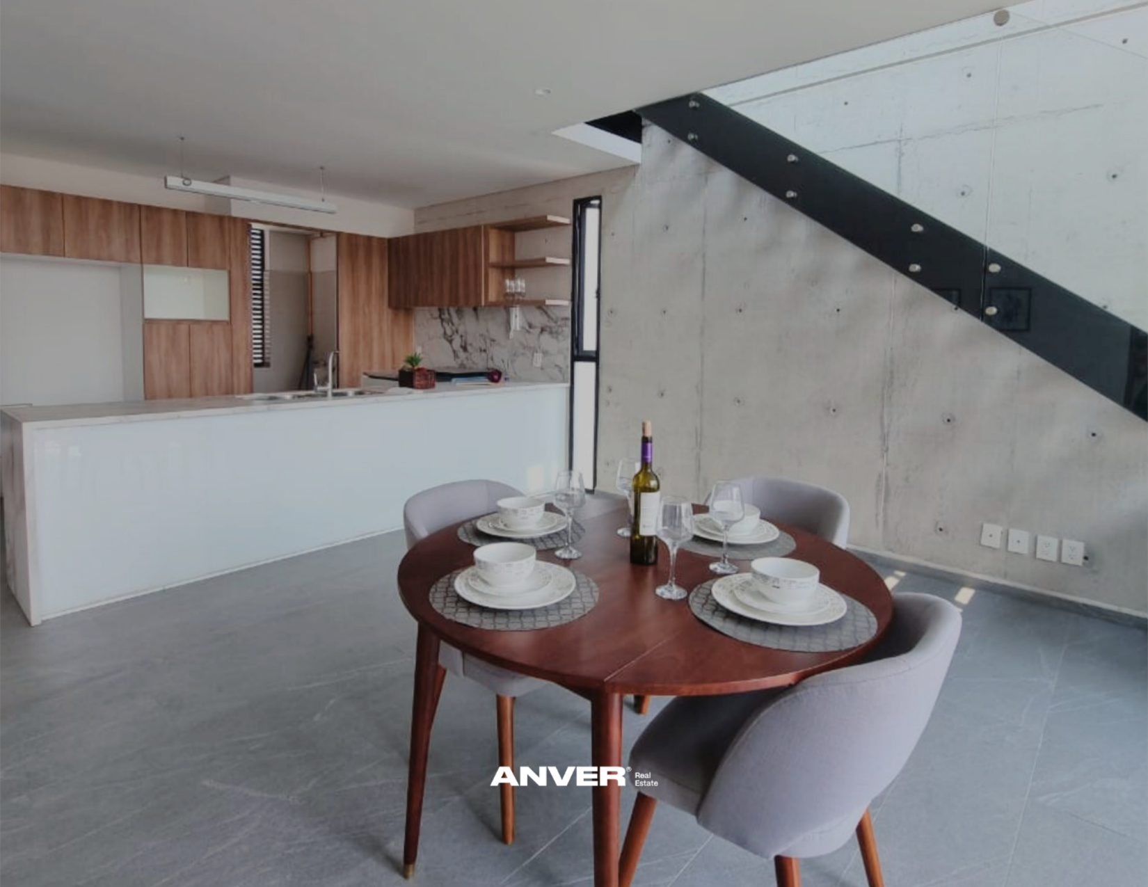 Interiores de una casa moderna con cocina y comedor, mesa redonda con platos, vasos y botella de vino, paredes de cemento y madera, escaleras de metal.