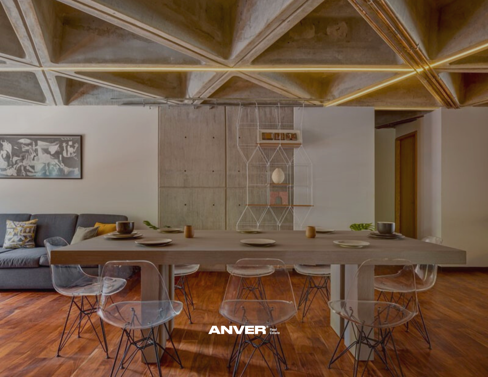 Sala de comedor moderna con mesa de madera, sillas de acrílico, un sofá gris con cojines decorativos, cuadro en la pared y repisas decorativas en la pared de fondo.
