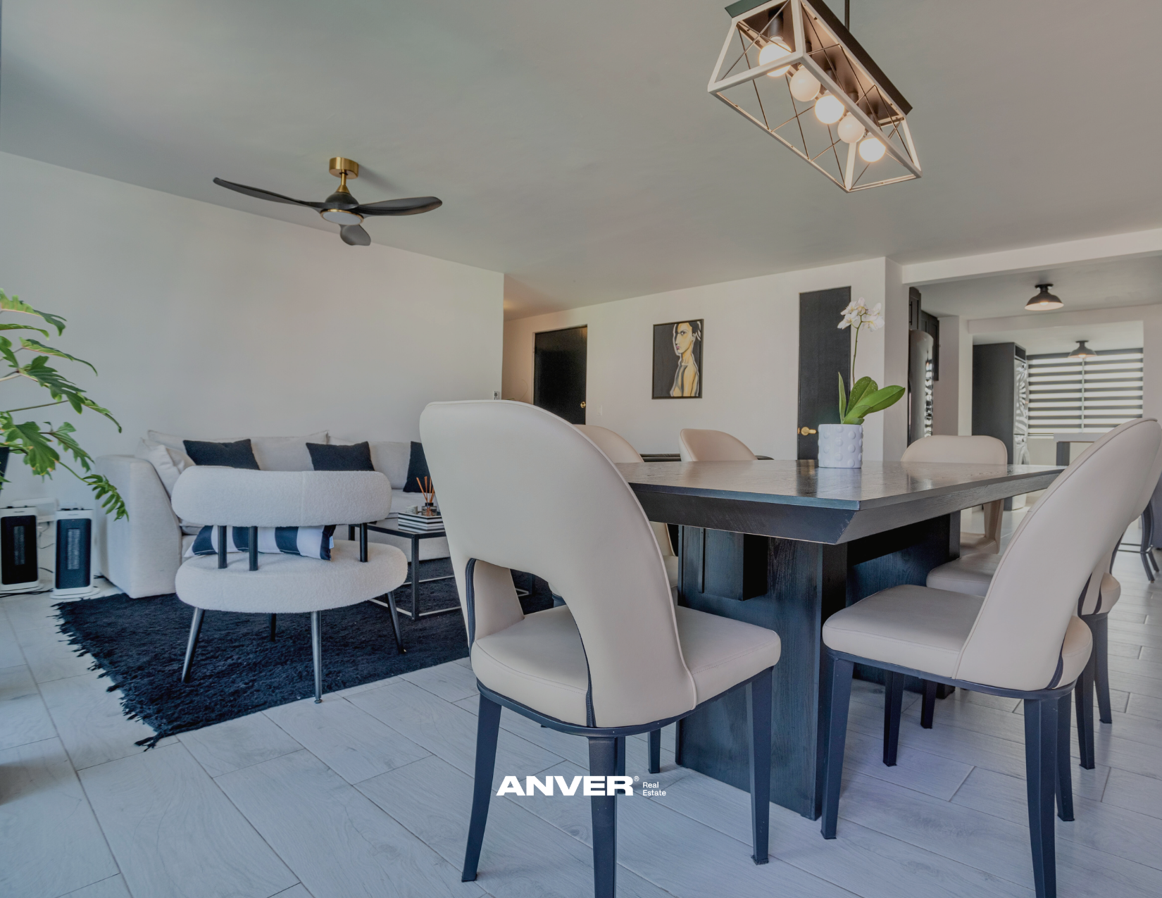 Sala de estar y comedor moderna con muebles en tonos claros y oscuros, una planta en una maceta blanca con flores blancas, cuadros en la pared, ventilador de techo negro y lámpara decorativa en el techo.