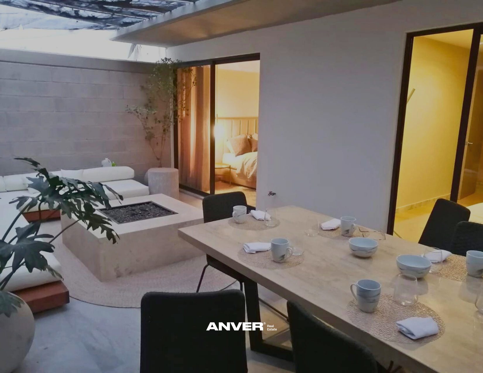 Comedor con mesa y sillas, en el fondo sala con sofá y cama, ventana y puerta en paredes blancas, plantas decorativas, iluminación cálida.
