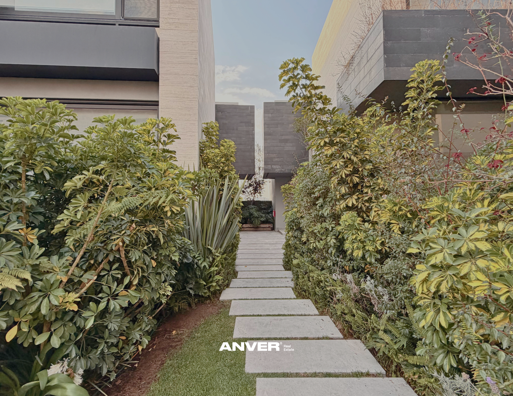 Camino de piedra en medio de un jardín con plantas verdes a cada lado, rodeado de edificios modernos con ventanas y paredes de piedra, con cielo claro en el fondo.