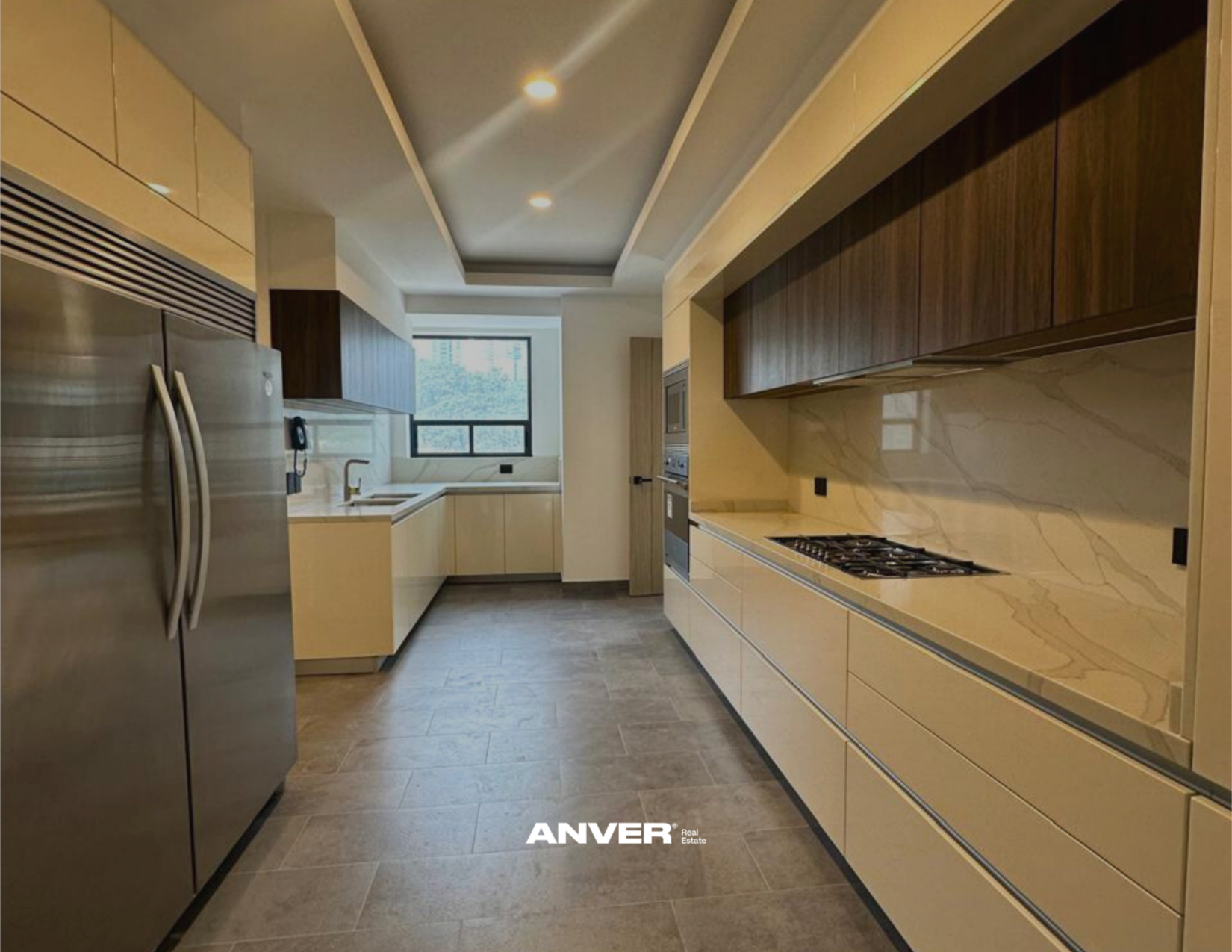 Cocina moderna con armarios de madera clara y encimeras de piedra beige, horno empotrado, estufa de gas y refrigerador de acero inoxidable, con ventana al exterior y piso de baldosa gris.