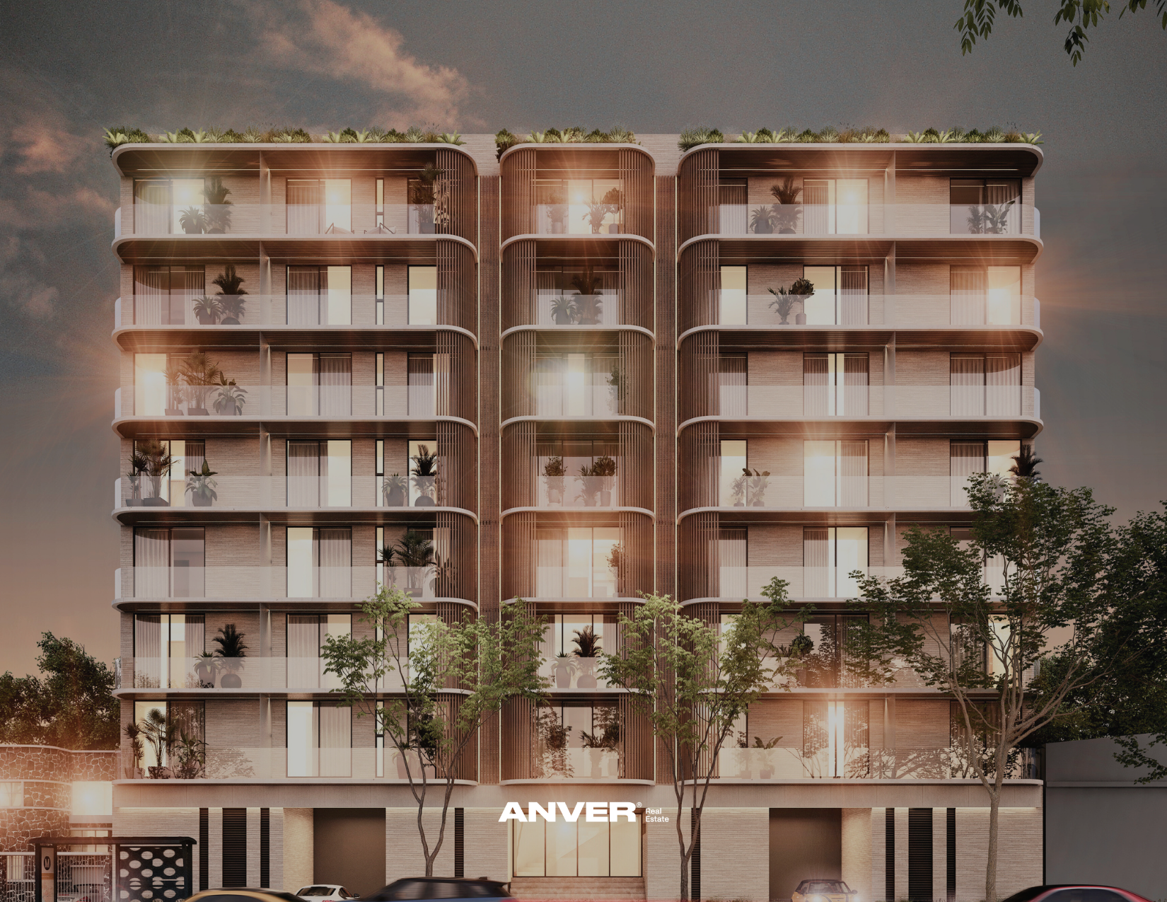 Edificio moderno de varios pisos con balcones y plantas en las terrazas, árboles en la calle y autos estacionados al frente, bajo un cielo al atardecer.
