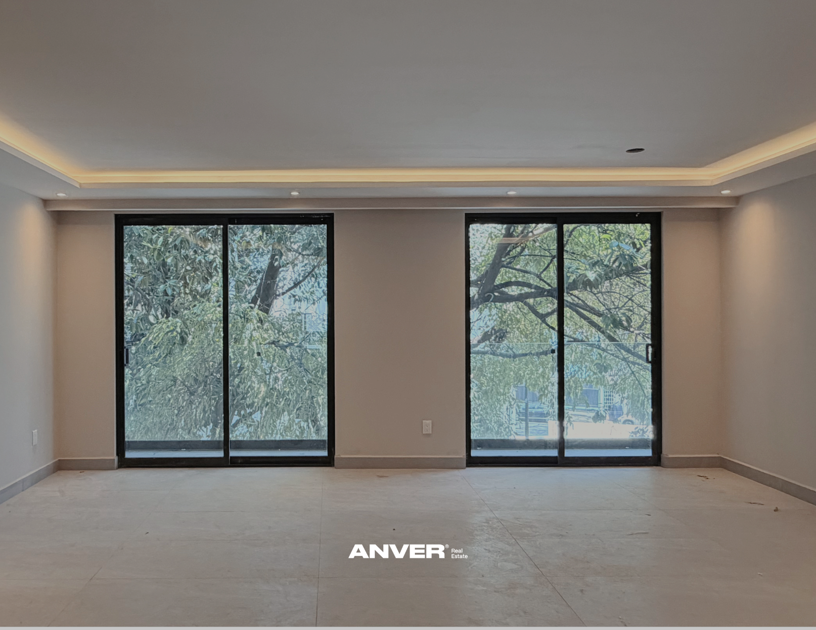 Interior de una sala con paredes blancas, ventanas grandes de vidrio con vista a árboles con hojas verdes, suelo de baldosas claras, techo con iluminación empotrada y moldura perimetral