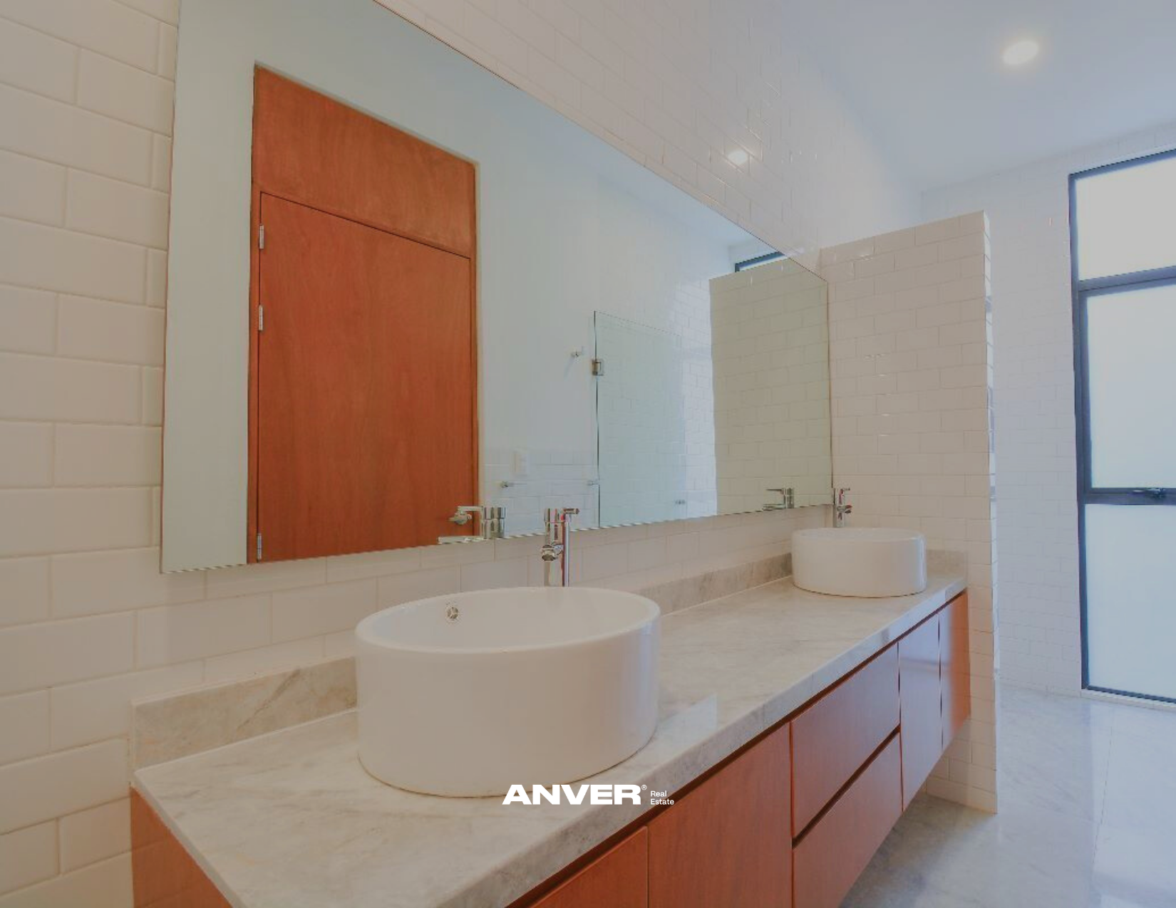 Lavabo y espejo en un baño con paredes de azulejo blanco y mueble de madera con encimera de piedra, con ventana grande y puerta de madera.