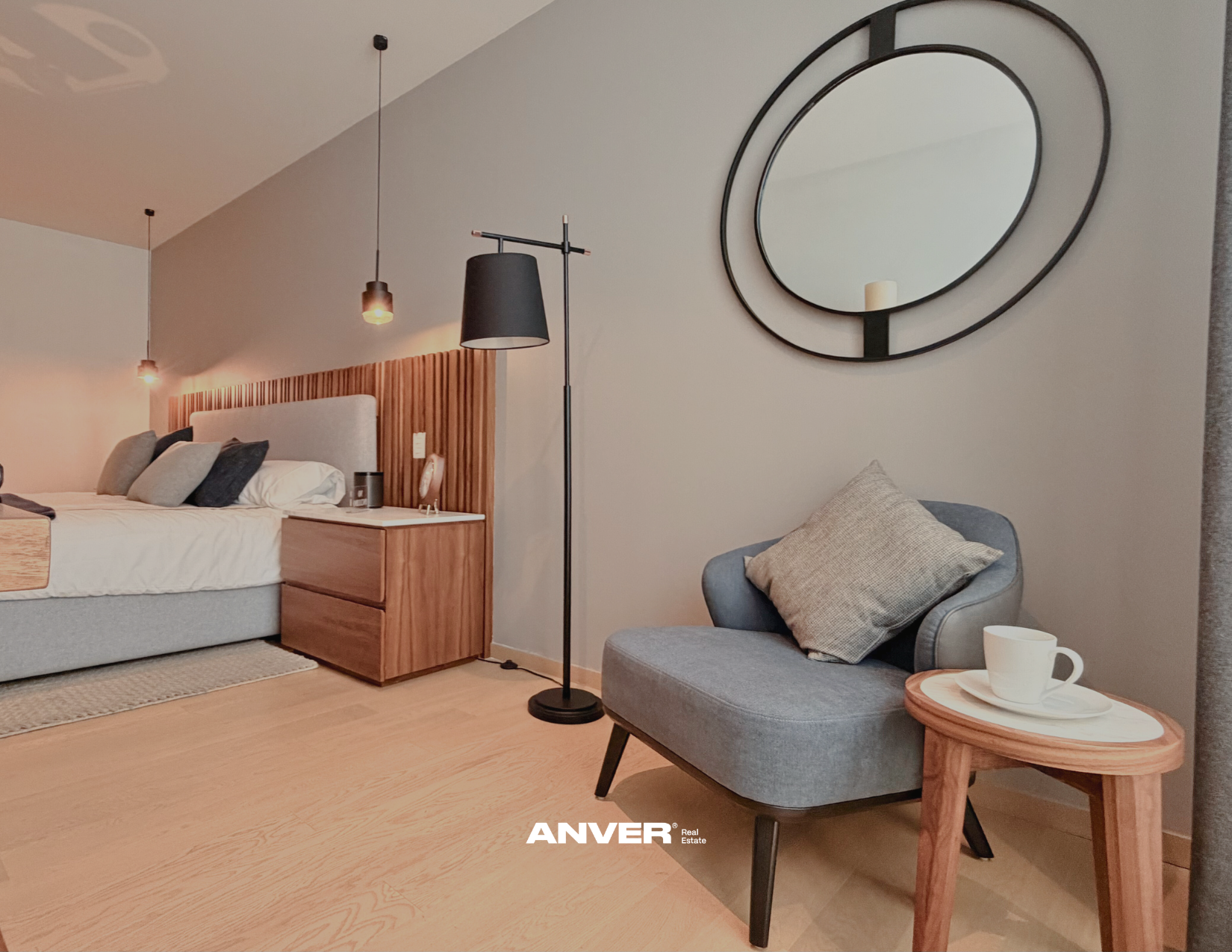 Sala moderna con cama, sillón gris, mesa de madera con taza y plato, espejo decorativo en la pared, lámparas colgantes y lámpara de pie.