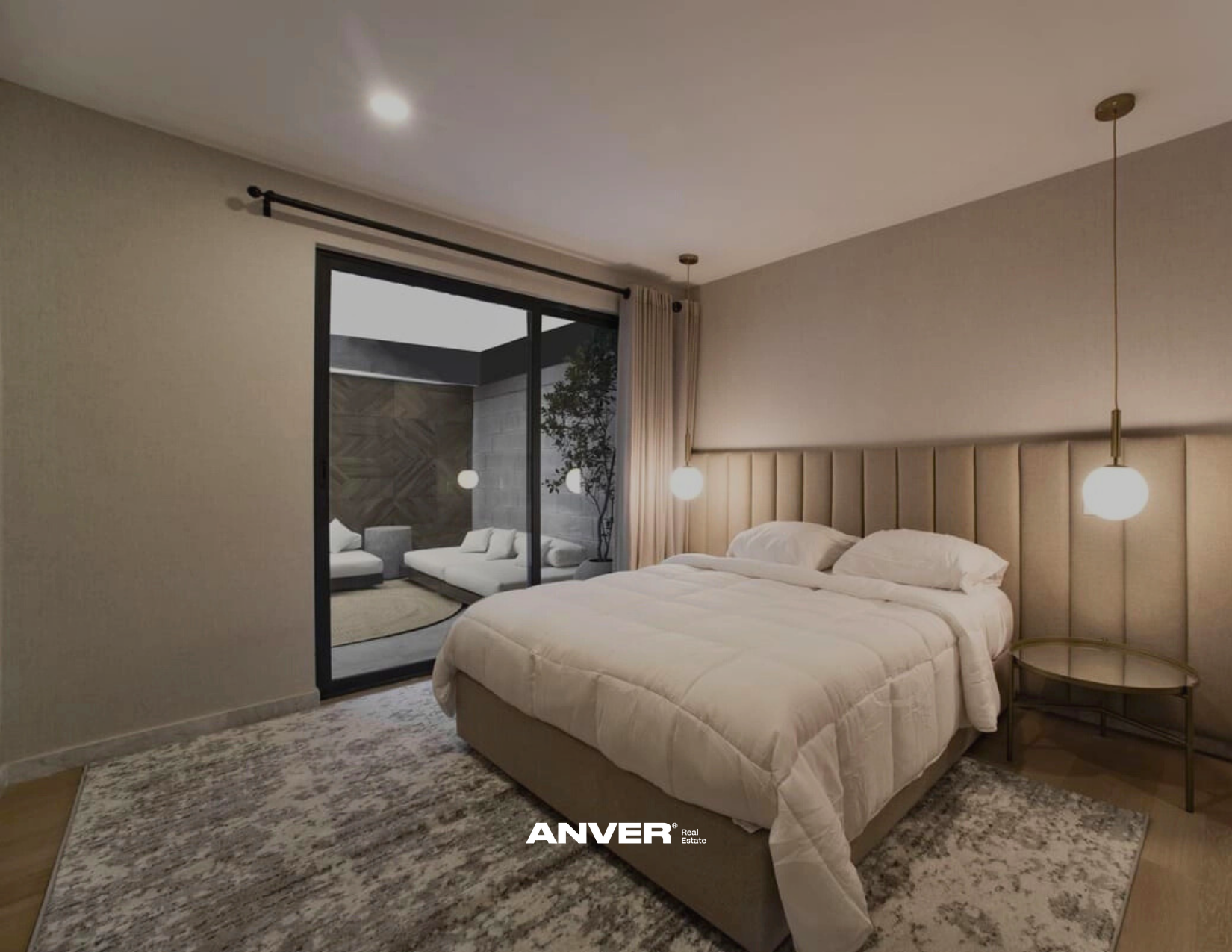 Habitación moderna con cama grande, cabecera acolchada, lámparas de suspensión y área de descanso exterior con sillones blancos y planta decorativa.