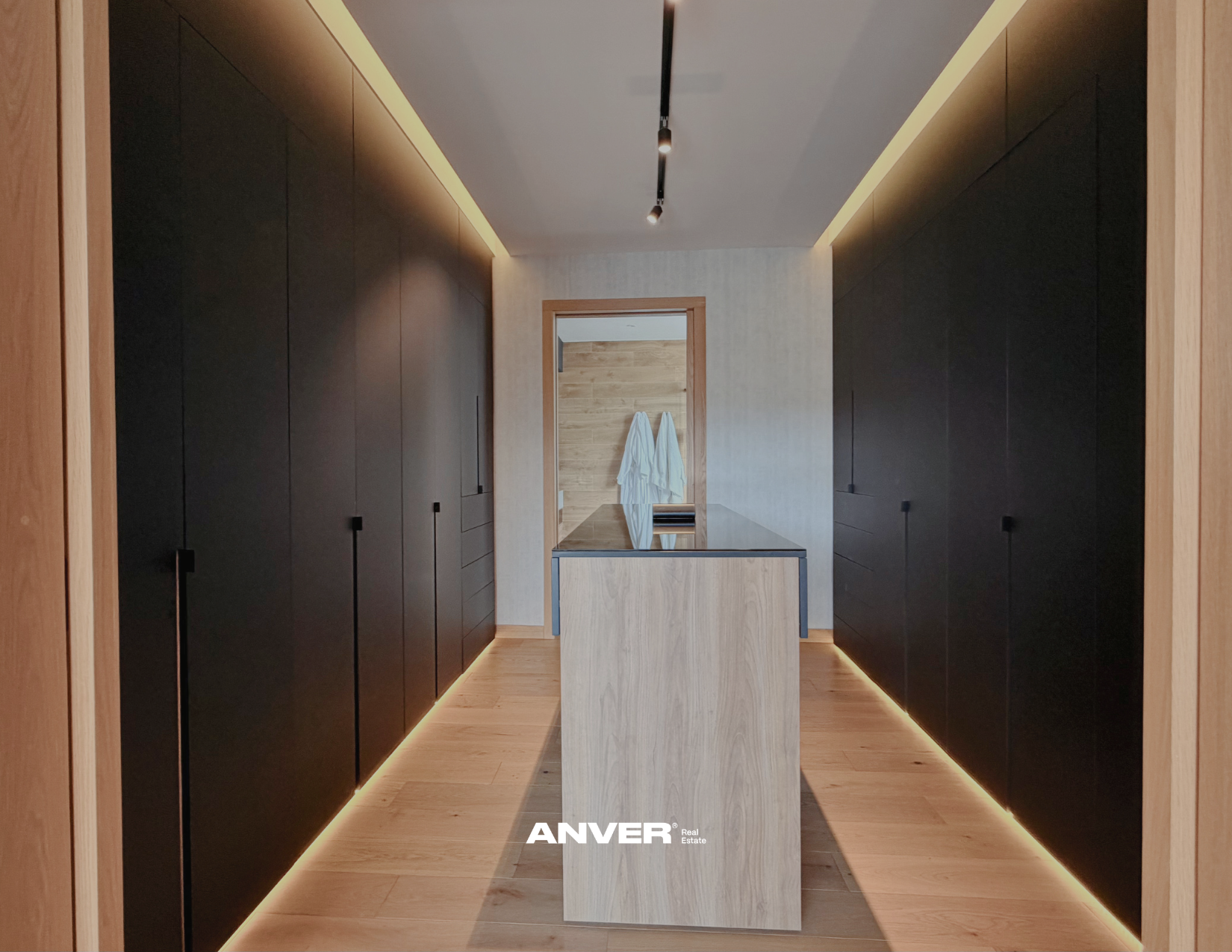 Vestidor moderno con armarios negros a ambos lados, una isla central de madera clara, iluminación Led en el techo y en el suelo, al fondo hay una puerta con toallas blancas colgadas.