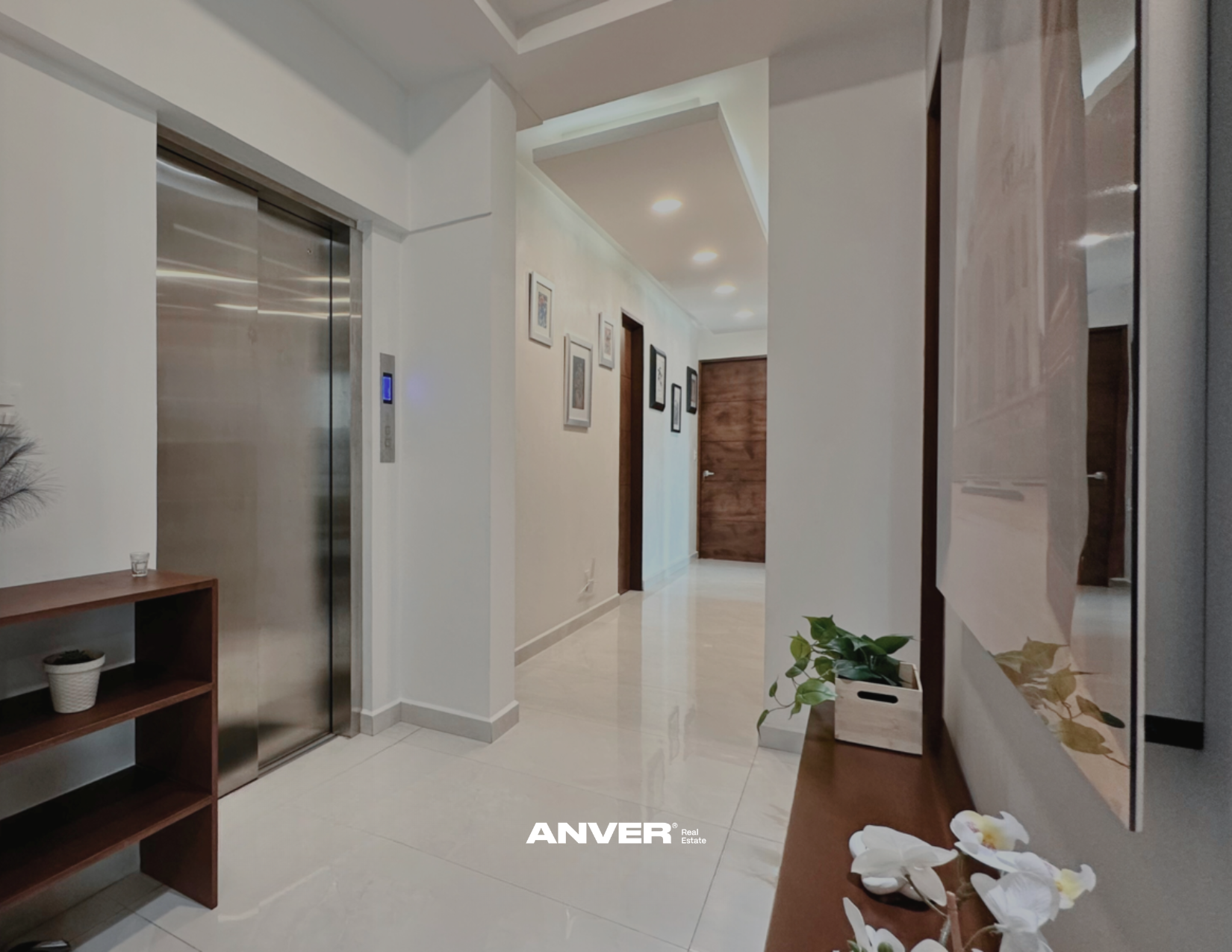 Vestíbulo con ascensor de acero inoxidable a la izquierda, pared blanca con cuadros en la pared del fondo y puerta de madera al centro, piso de baldosas blancas, plantas decorativas y mirador en la pared derecha, iluminación empotrada.