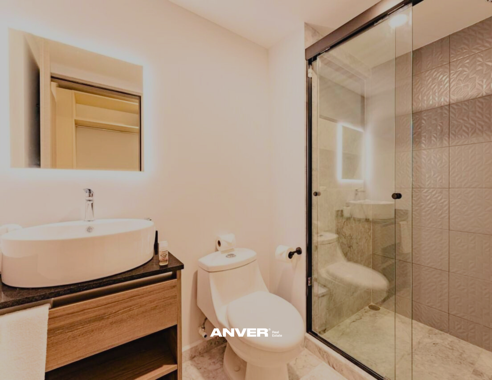 Baño moderno con lavabo ovalado en mueble de madera, espejo con luz, inodoro, papel higiénico, ducha con puertas de vidrio y paredes decoradas con azulejos texturizados.