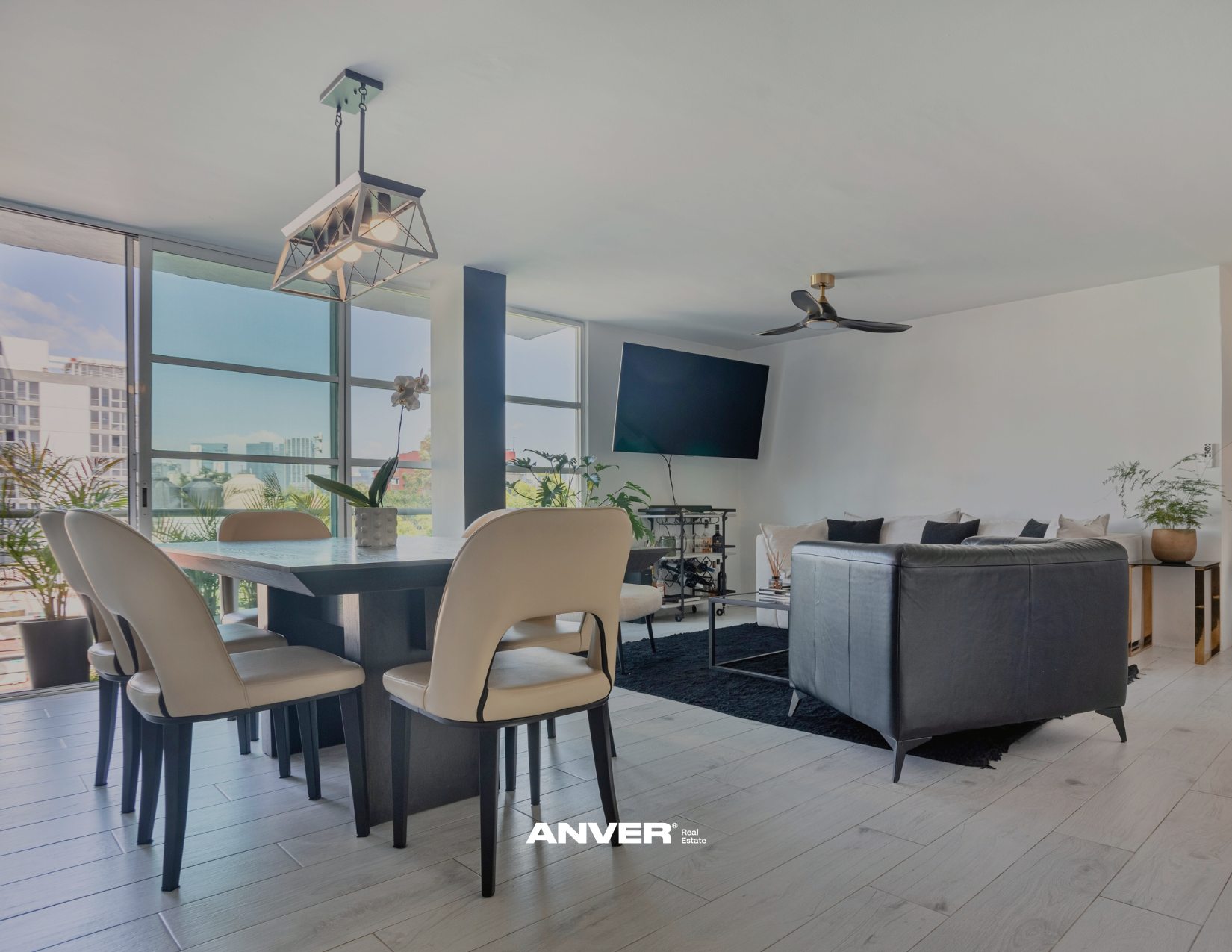 Sala de estar y comedor en apartamento con ventanas grandes, plantas, televisor en pared, sofá gris y comedor con sillas blancas.