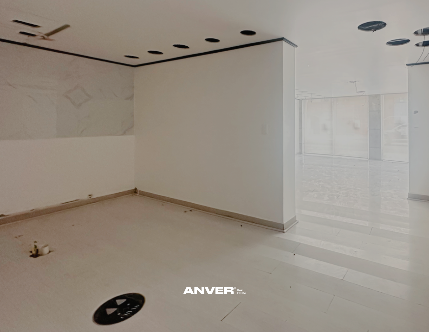 Interior de un espacio en construcción o remodelación, con paredes blancas, piso de cerámica y accesorios de plomería en el suelo y en la pared, con ventanas grandes que dejan entrar luz natural.