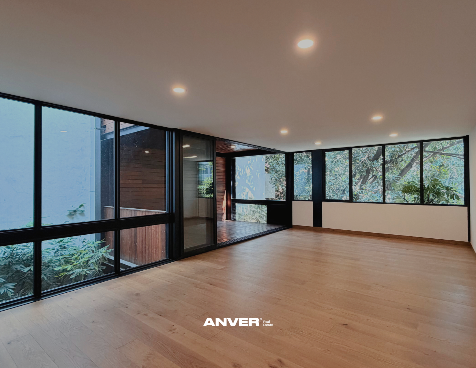 Interior de una sala moderna con ventanas grandes y plantas visibles afuera, suelo de madera y techo con luces empotradas.