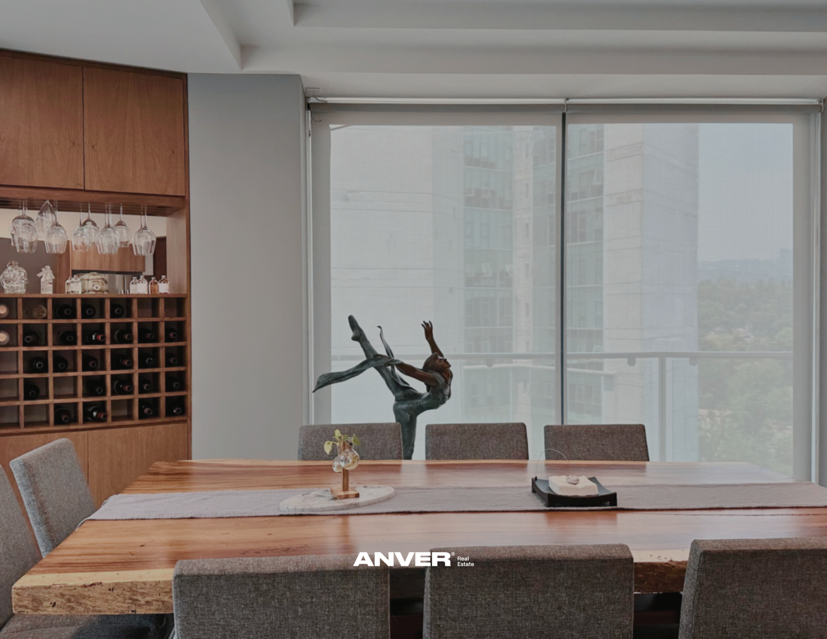 Una mesa de comedor de madera con seis sillas tapizadas en tela gris claro. En el centro, una pequeña planta en maceta y una figura decorativa de una bailarina. En el fondo, una pared con una vitrina de vino y cristalería, y ventanas grandes con vista a edificios de la ciudad.
