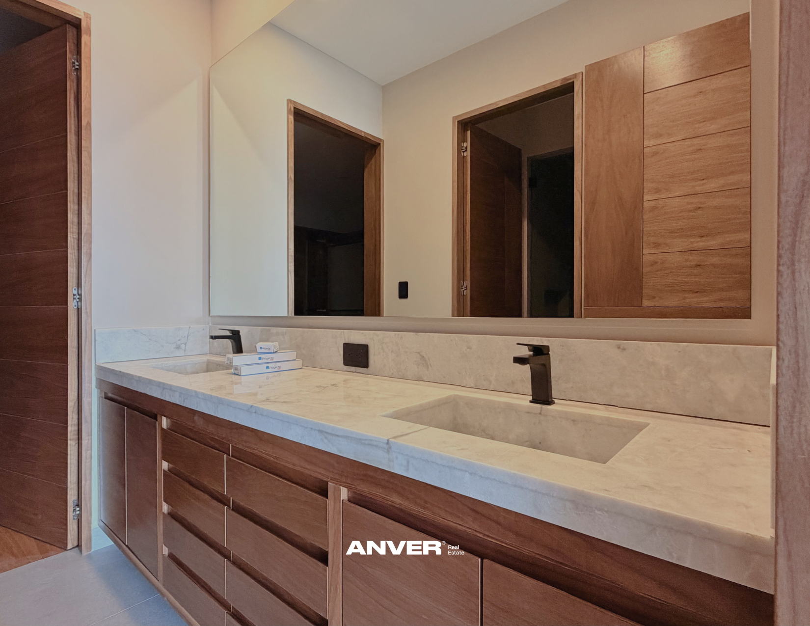 Lavabo con encimera de mármol, grifos negros, espejo grande y armarios de madera en un baño moderno.