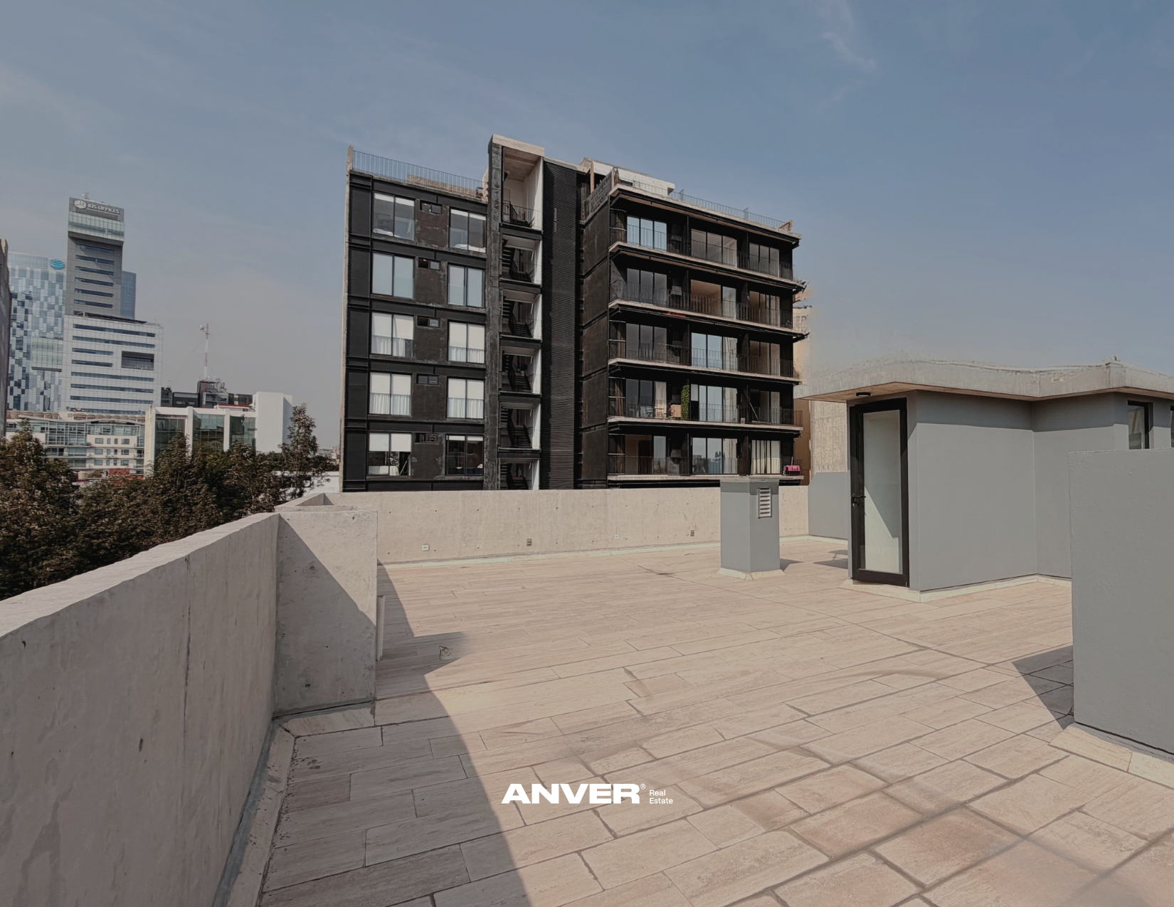 Vista de una azotea con suelo de baldosas y una estructura pequeña en un lado, con edificios altos al fondo, uno de ellos de color negro con balcones y ventanas grandes, y otros edificios en la ciudad.