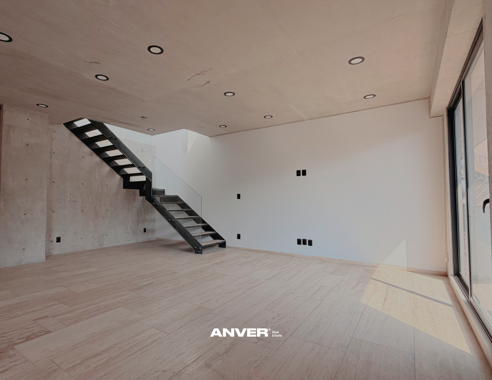 Espacio interior moderno con paredes de concreto y blanco, escalera negra con barandal de vidrio, pisos de madera clara, ventanas de vidrio grandes y luces empotradas en el techo.