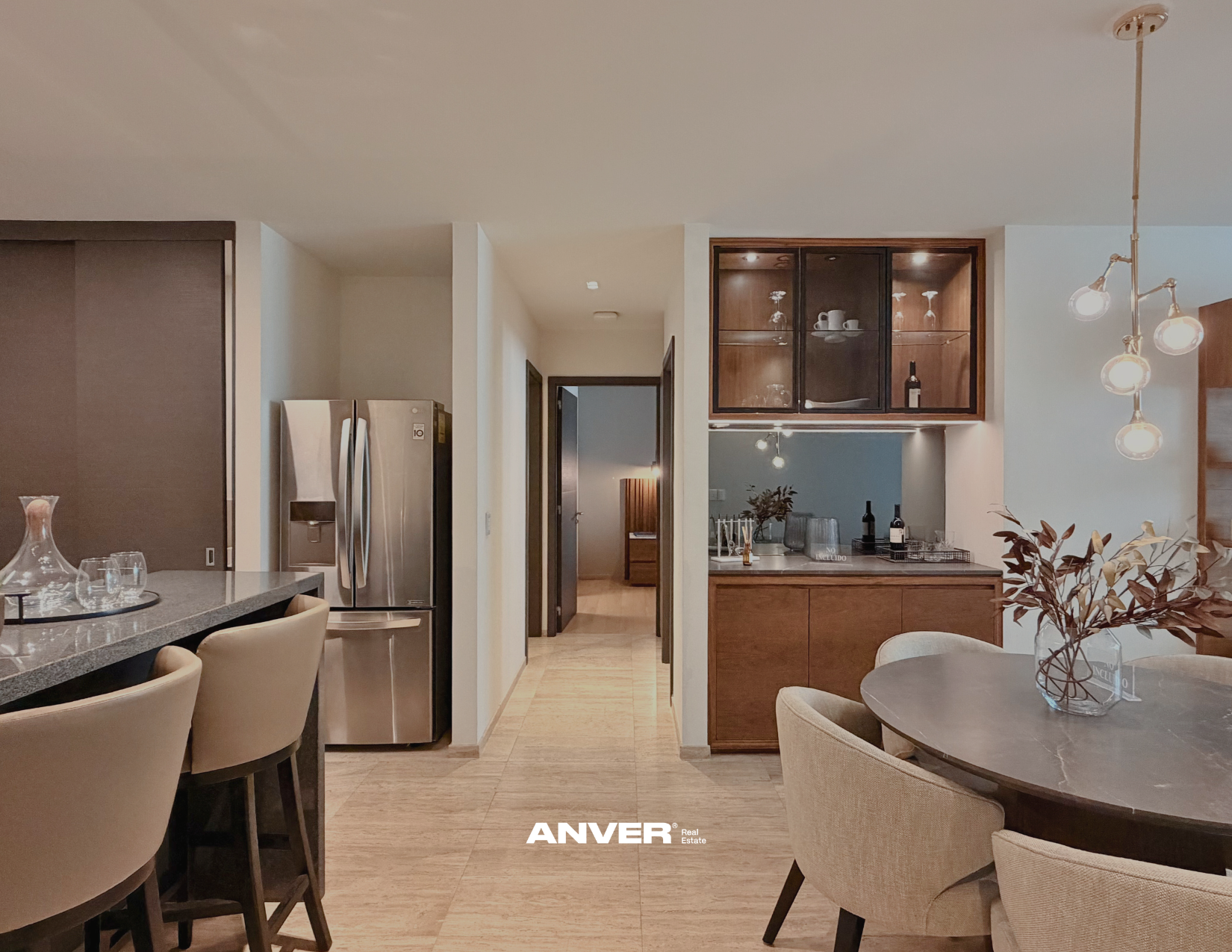 Vista de una sala de estar y comedor en una casa moderna con mesa redonda, sillas beige, plantas decorativas y una pared con gabinetes de madera. Incluye una cocina con nevera de acero inoxidable y una entrada hacia un pasillo.