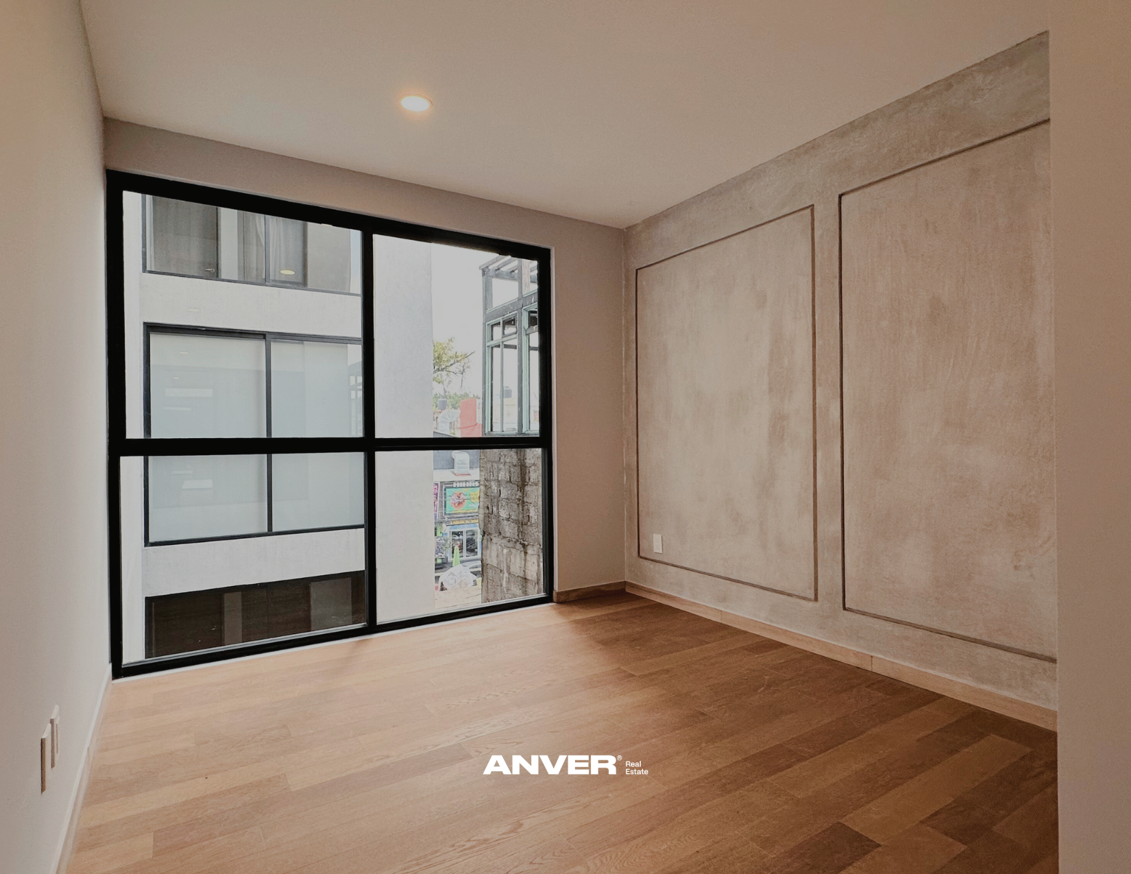 Habitación vacía con suelo de madera, paredes beige, y una ventana grande con marco negro que da a un exterior con edificios cercanos.