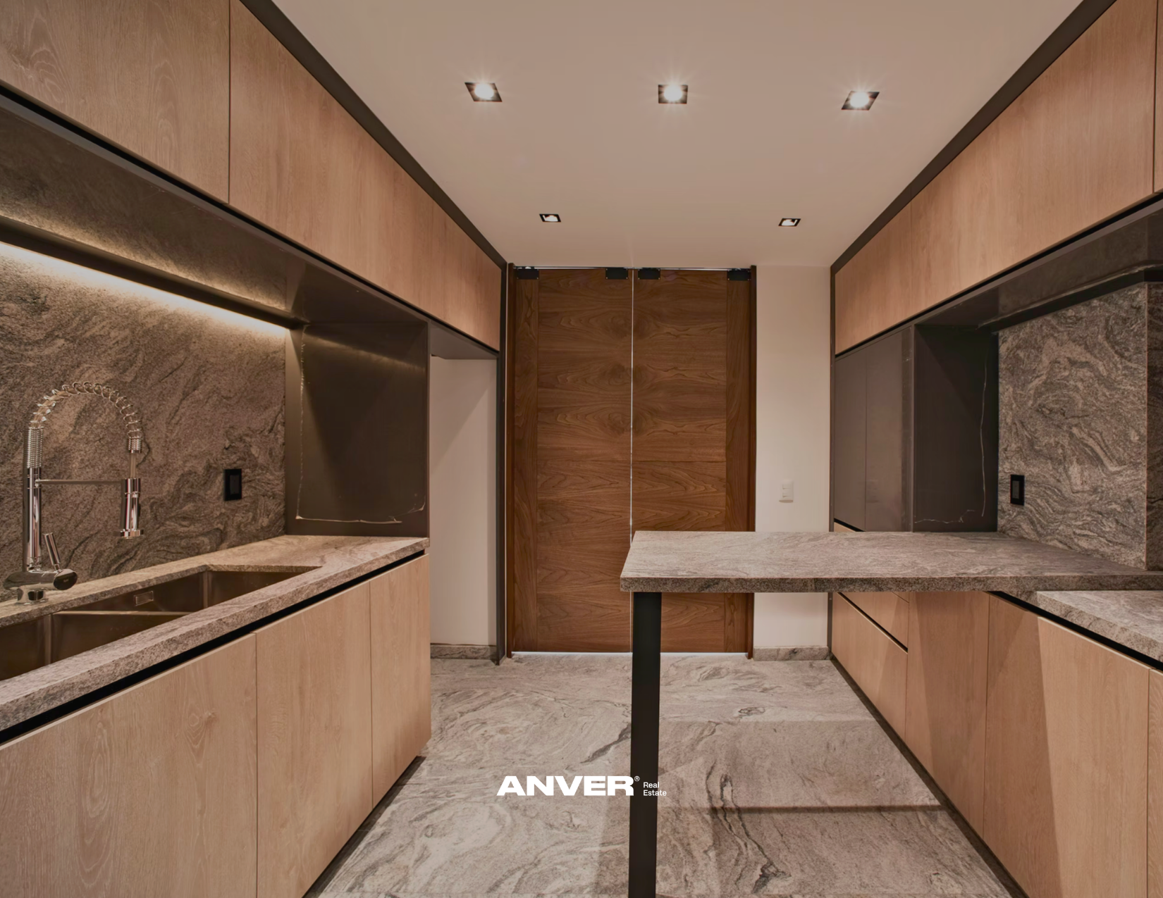 Cocina moderna con gabinetes de madera clara, encimera de granito, fregadero de acero inoxidable y paredes de piedra natural. Techo con luces empotradas y puerta de madera en el fondo.