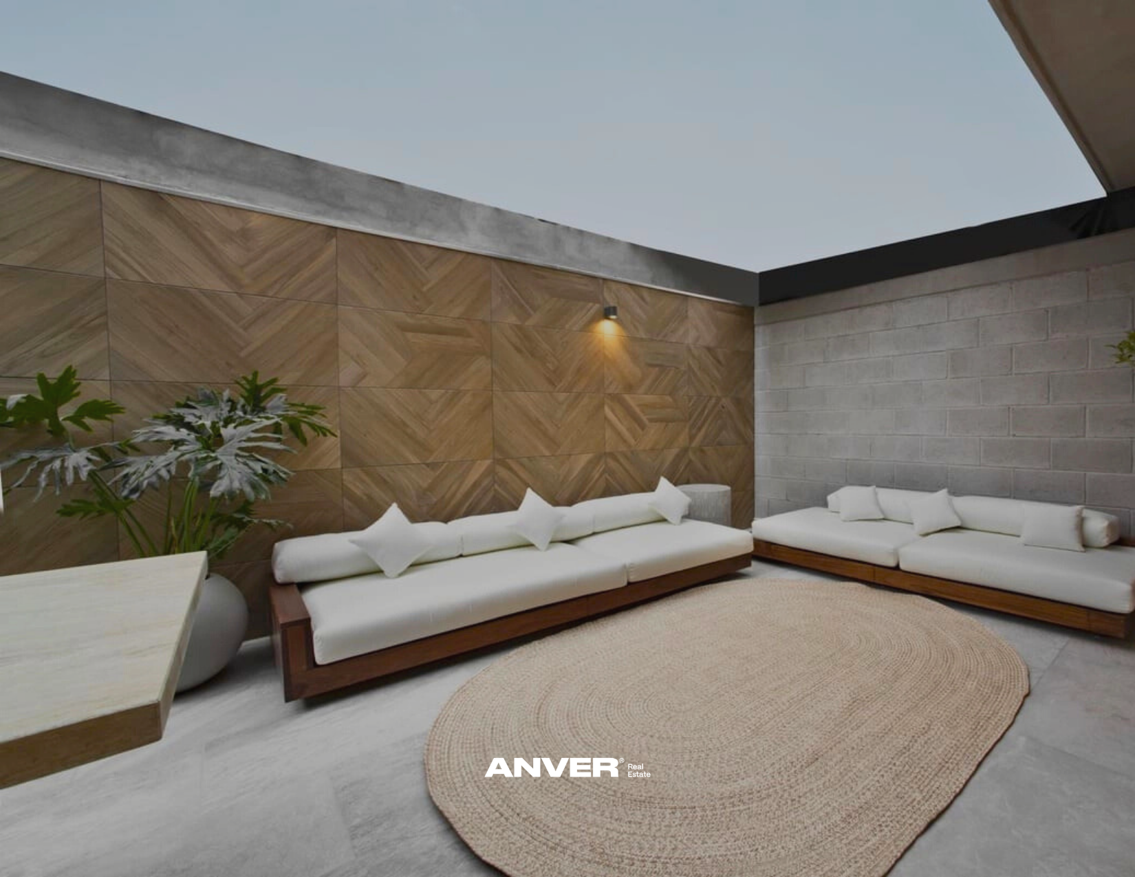 Sala techada con paredes de piedra y madera, dos sofás blancos con cojines, alfombra redonda de fibra, planta decorativa y luz de pared.