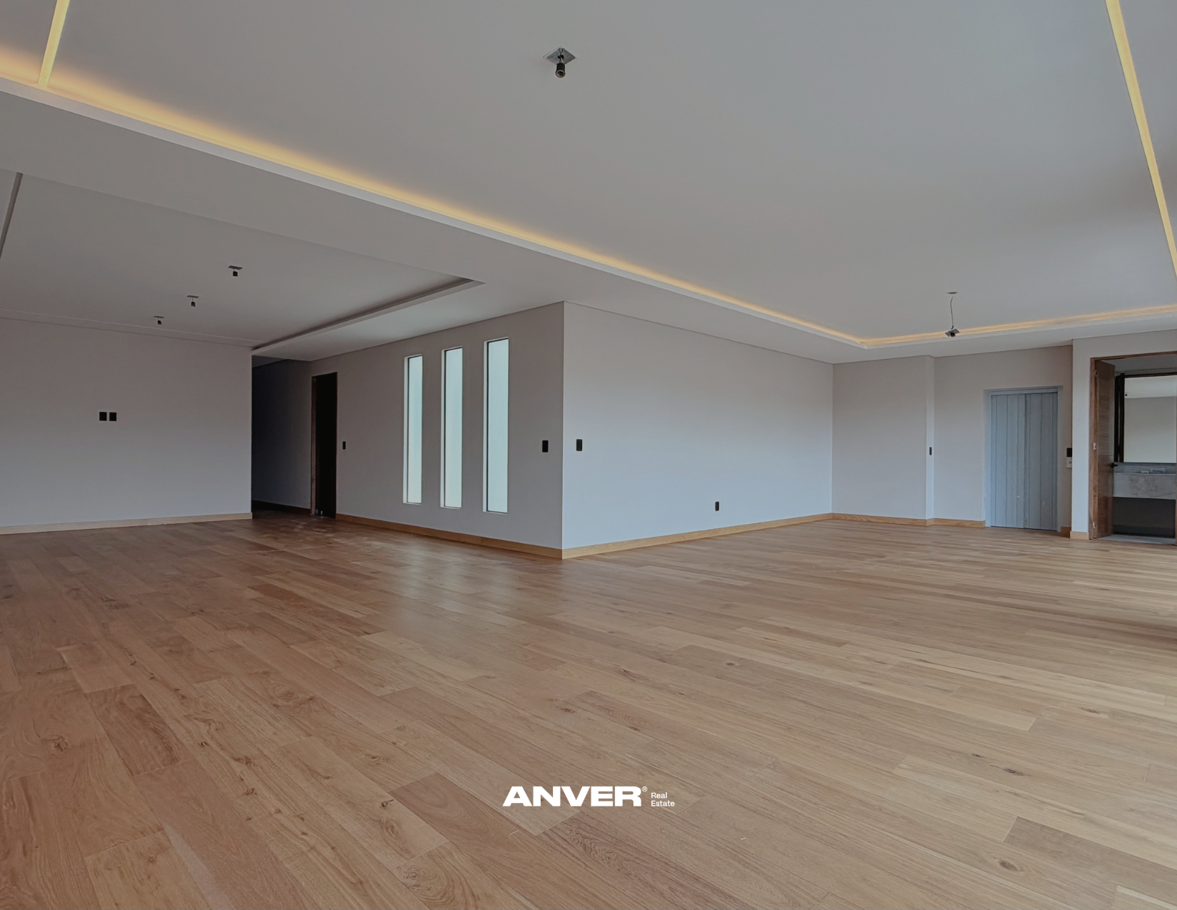 Interior vacío de una sala moderna con paredes blancas, ventanas estrechas y piso de madera clara.