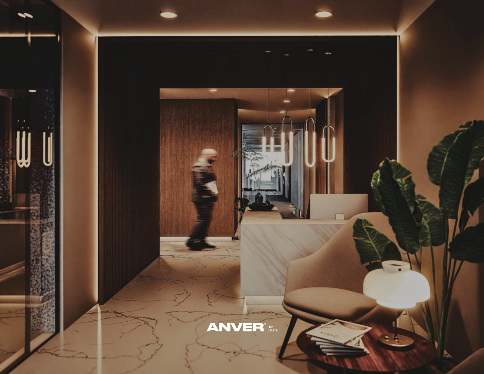Interior moderno con iluminación cálida, plantas verdes, mobiliario de recepción con escritorio de mármol y sillón beige, y un hombre caminando por un pasillo.