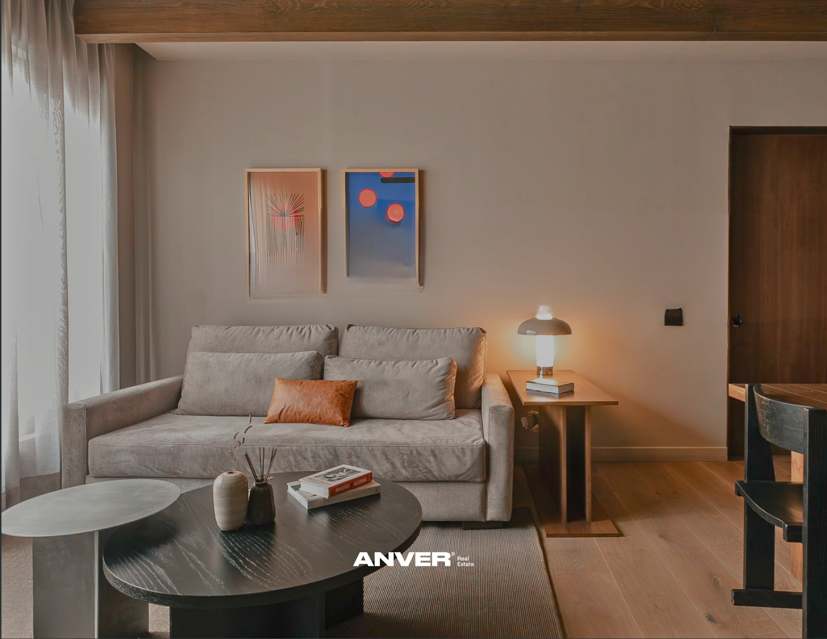 Salón de una sala de estar con sofá beige, mesita de centro con libros y decoraciones, lámpara encendida, cuadros modernos en la pared y cortinas blancas que dejan entrar luz natural.