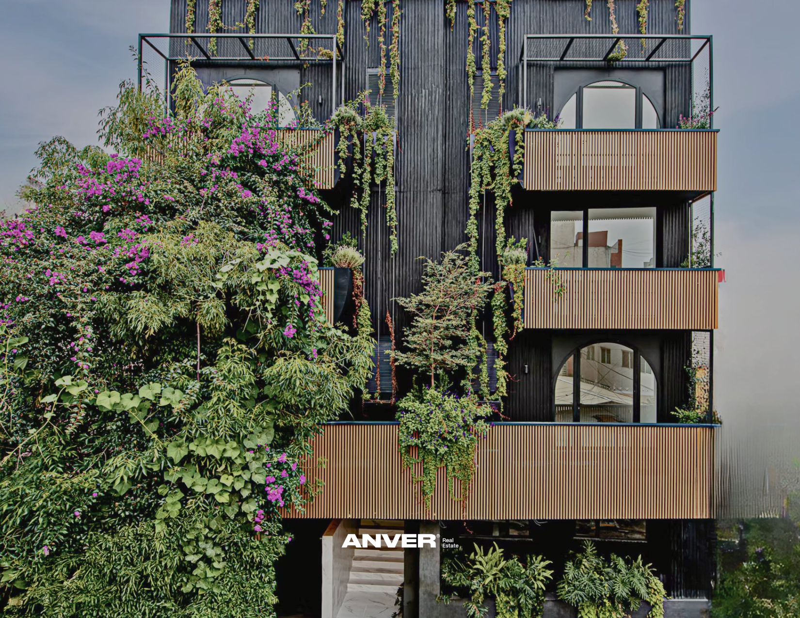 Edificio moderno de varios pisos con rejas de madera en los balcones y paredes negras, rodeado de vegetación con arbustos y enredaderas con flores violetas y verdes