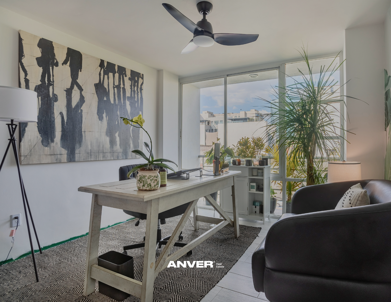 Sala de estar con mesa de madera, plantas, sillón negro, pared con pintura abstracta y ventanas grandes con vista a edificios urbanos y cielo nublado.