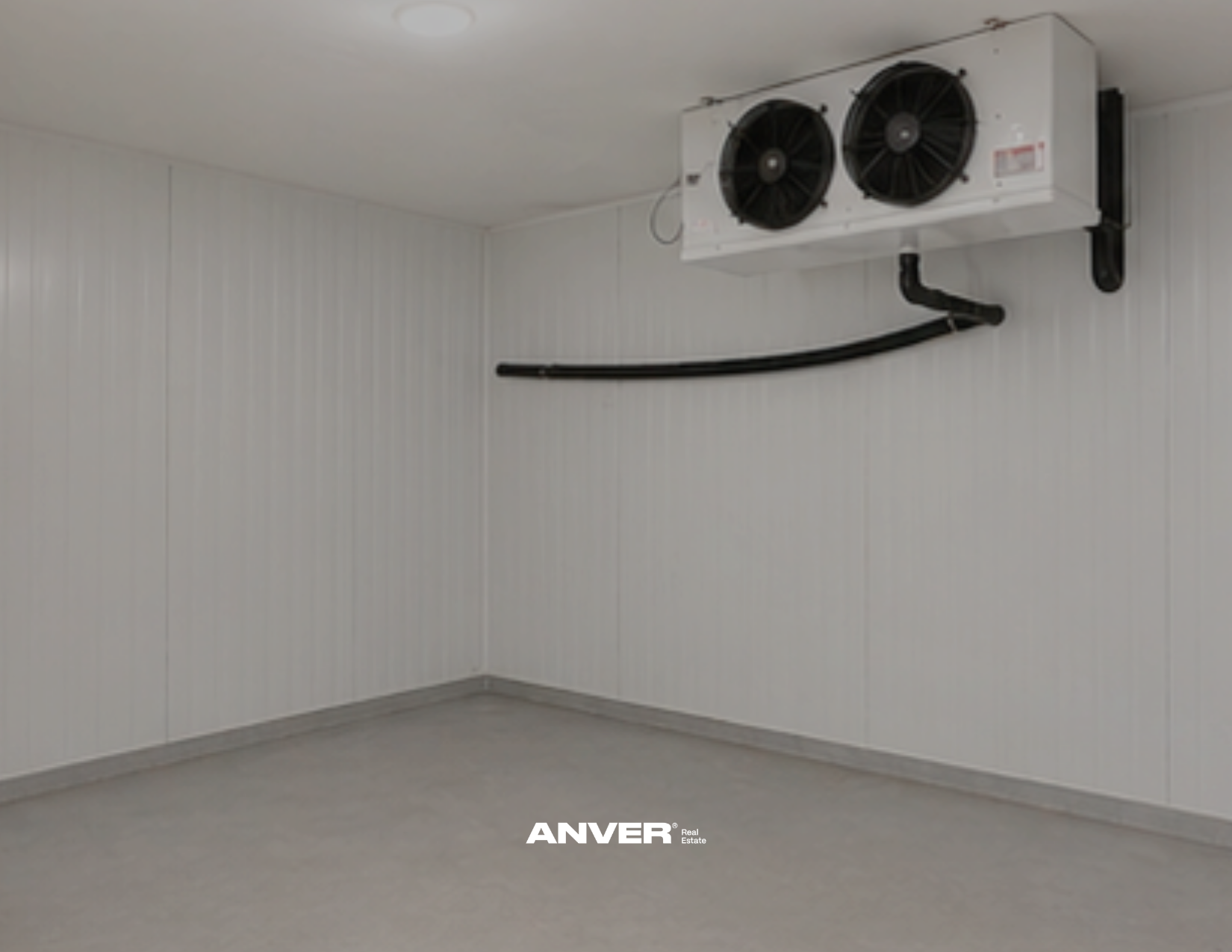 Paredes blancas y suelo de color gris en una habitación vacía. En la parte superior, un sistema de refrigeración con dos ventiladores grandes y un tubo negro. En la parte inferior, el logo de "ANVER".